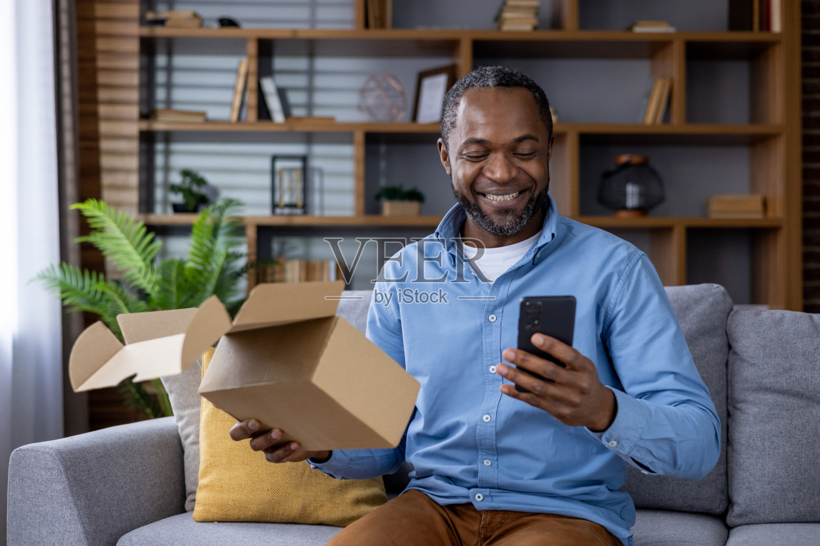 一名男子坐在家里的沙发上玩智能手机，享受打开快递包裹的乐趣照片摄影图片