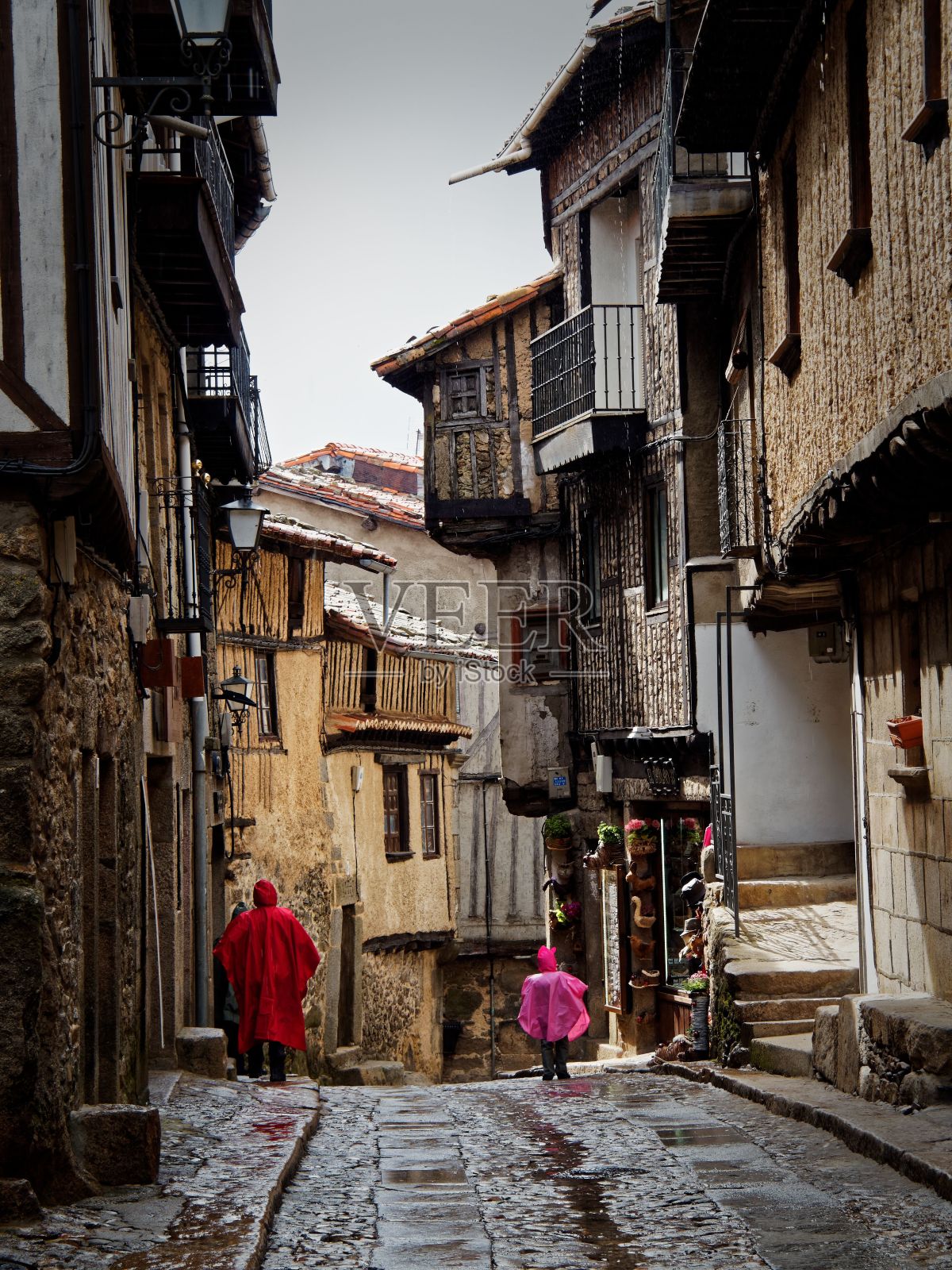 狭窄的鹅卵石街道在一个历史悠久的西班牙村庄,人们穿着五颜六色的雨衣照片摄影图片