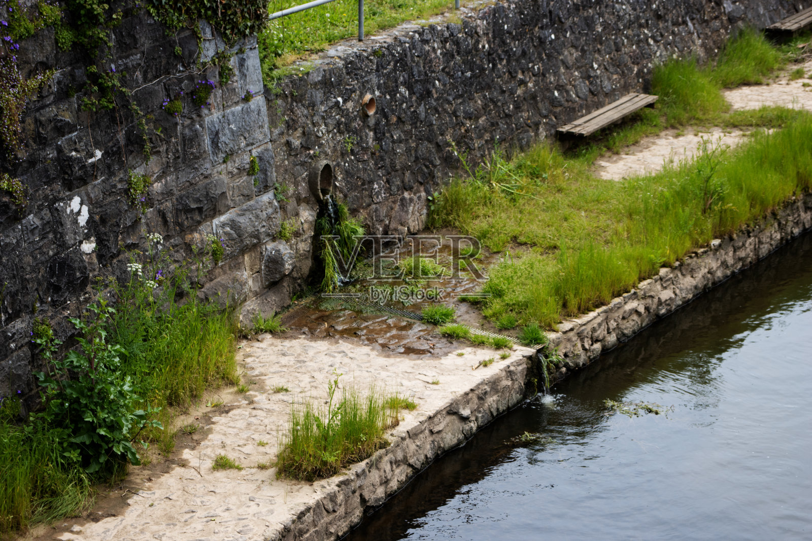 老旧肮脏的砖墙上有地衣和污垢，排水口流入流动的溪流照片摄影图片