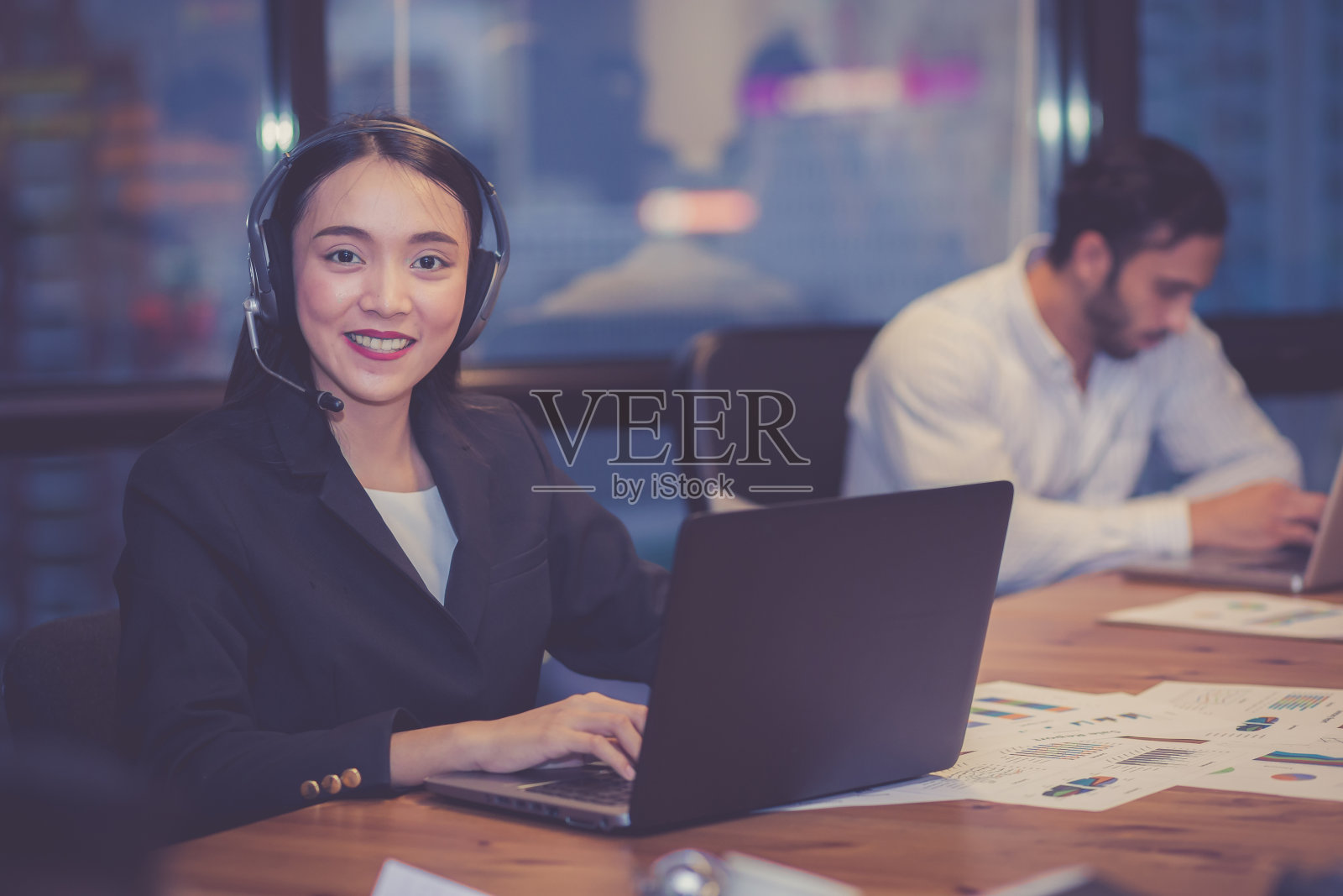 专业年轻的亚洲女商人戴着耳机在现代办公室里用笔记本电脑工作，同事在后台，女商人戴着耳机为客户服务，经营理念。照片摄影图片