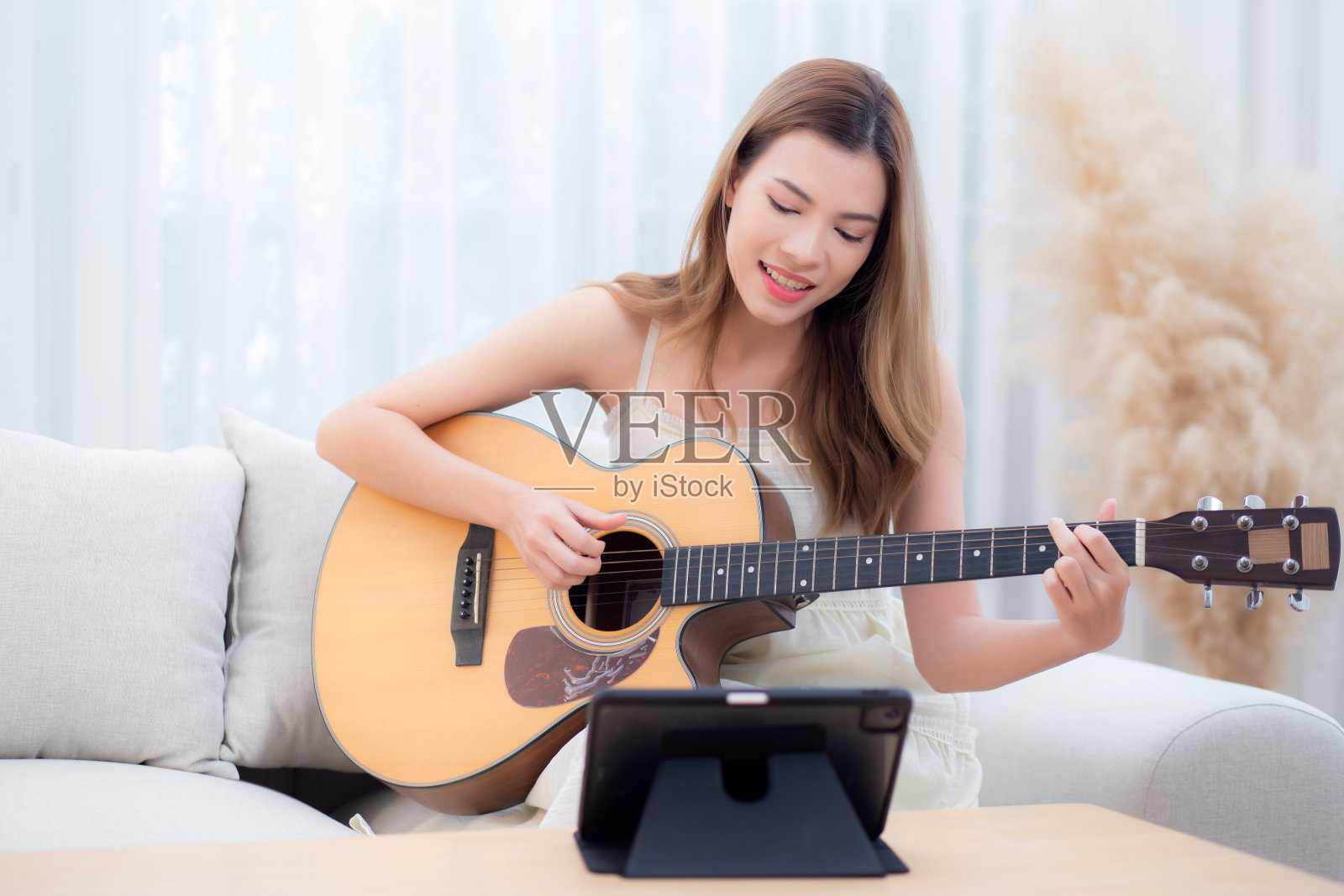 年轻的亚洲妇女坐在沙发上寻找平板电脑在家里的客厅里用吉他练习音乐，妇女在网上观看有关音乐和学习吉他的课程，生活方式概念。照片摄影图片