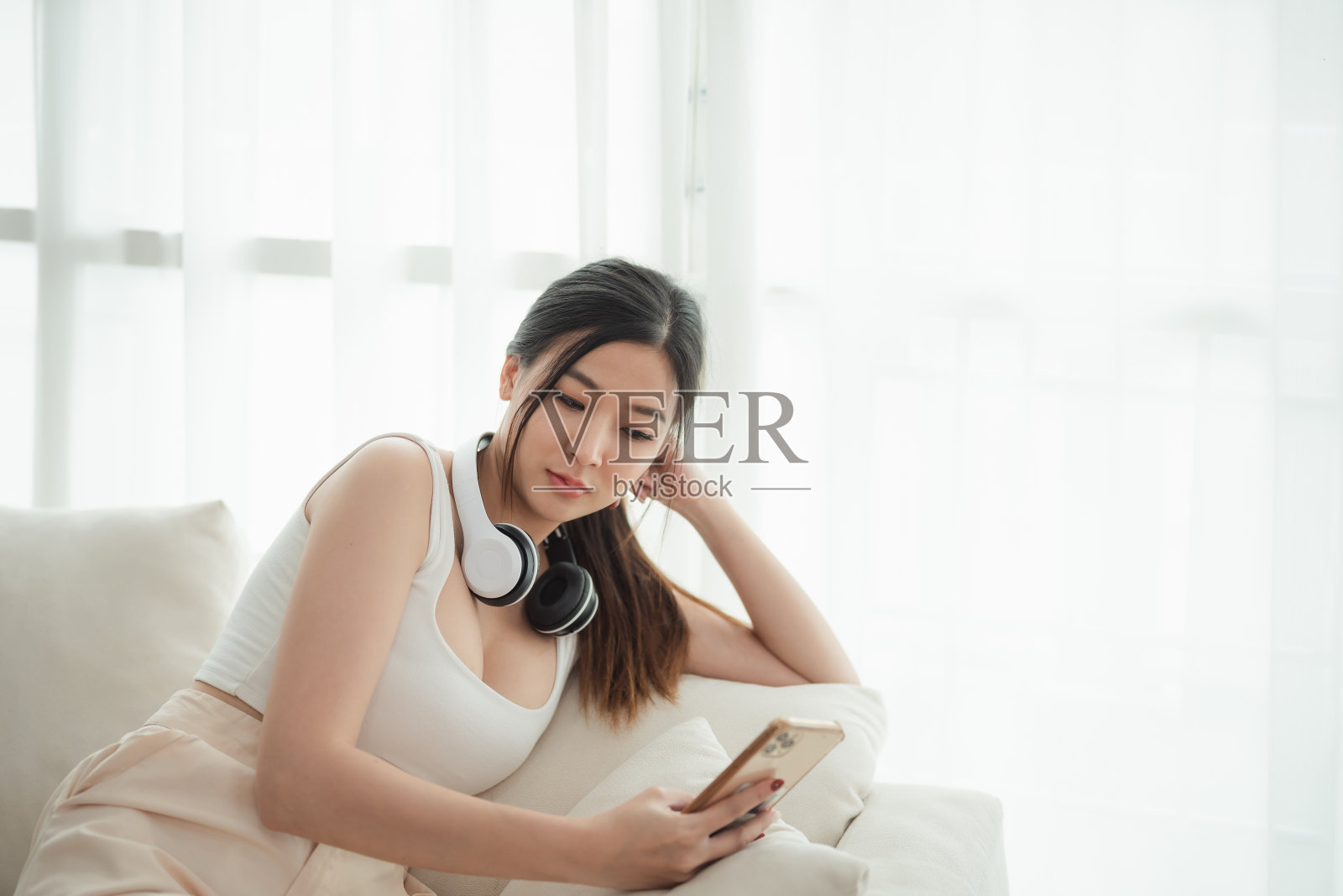 一个亚洲女人正坐在沙发上看手机。她戴着耳机，正在听音乐照片摄影图片
