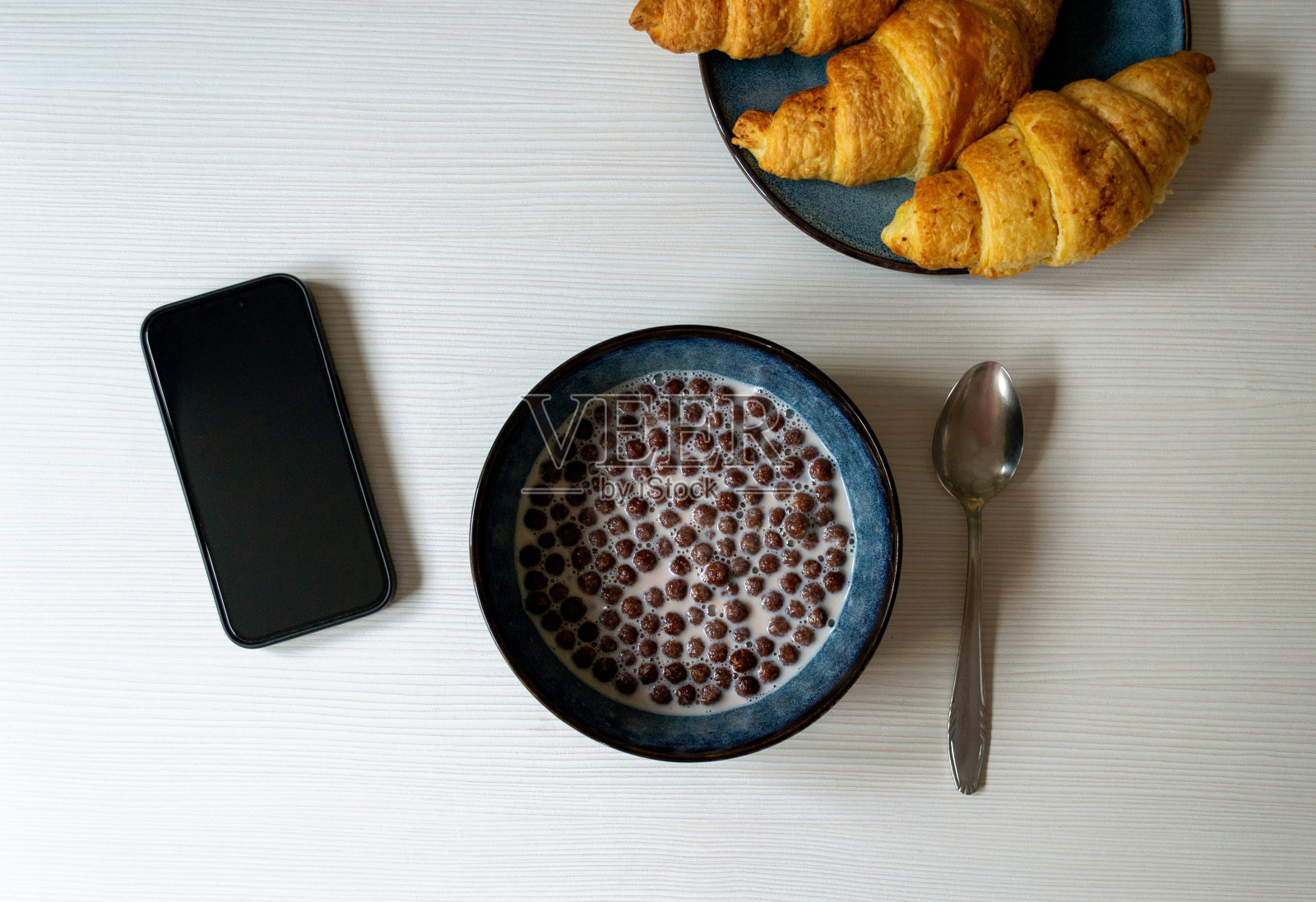 早餐:麦片、牛奶巧克力球、羊角面包和智能手机平放在桌上照片摄影图片