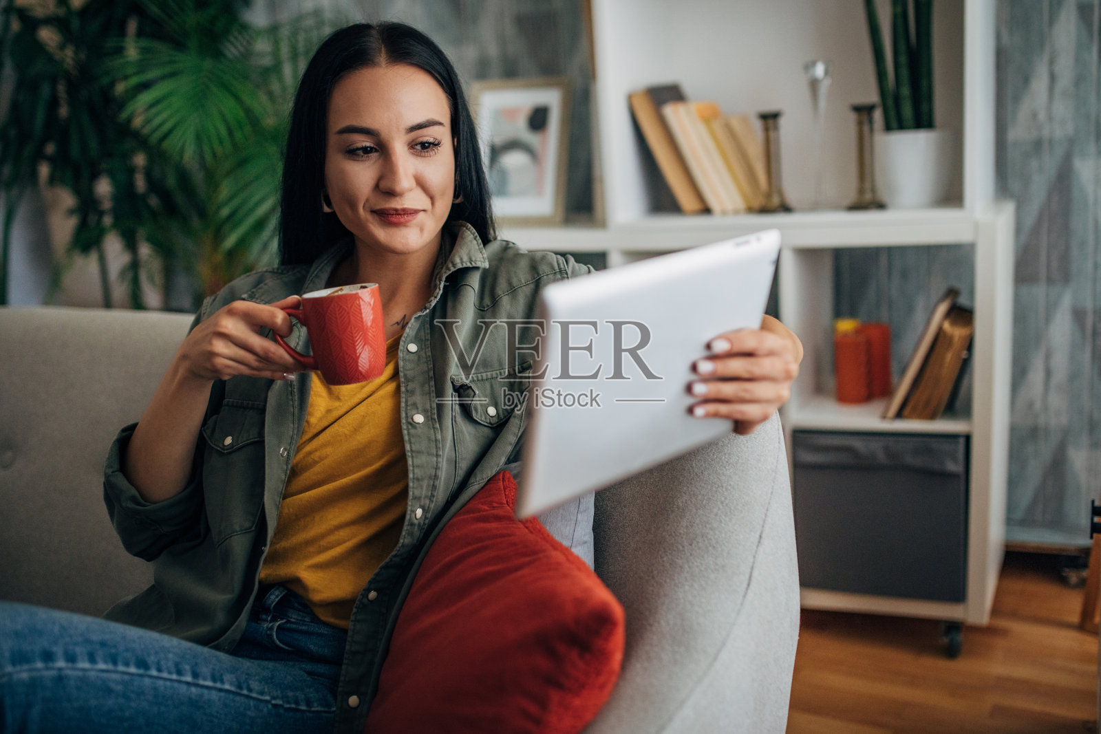 女人一边在沙发上浏览平板电脑一边喝咖啡。照片摄影图片