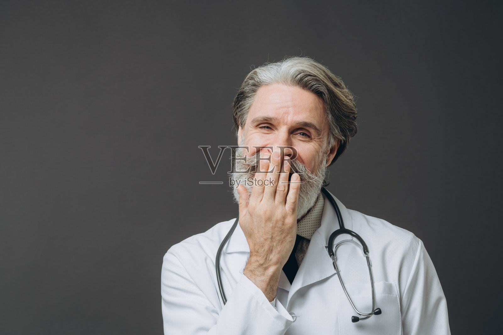 白发苍苍、身穿白大褂的男医生，在灰色的背景中孤立无援，双手捂着嘴，做着沉默的手势。照片摄影图片