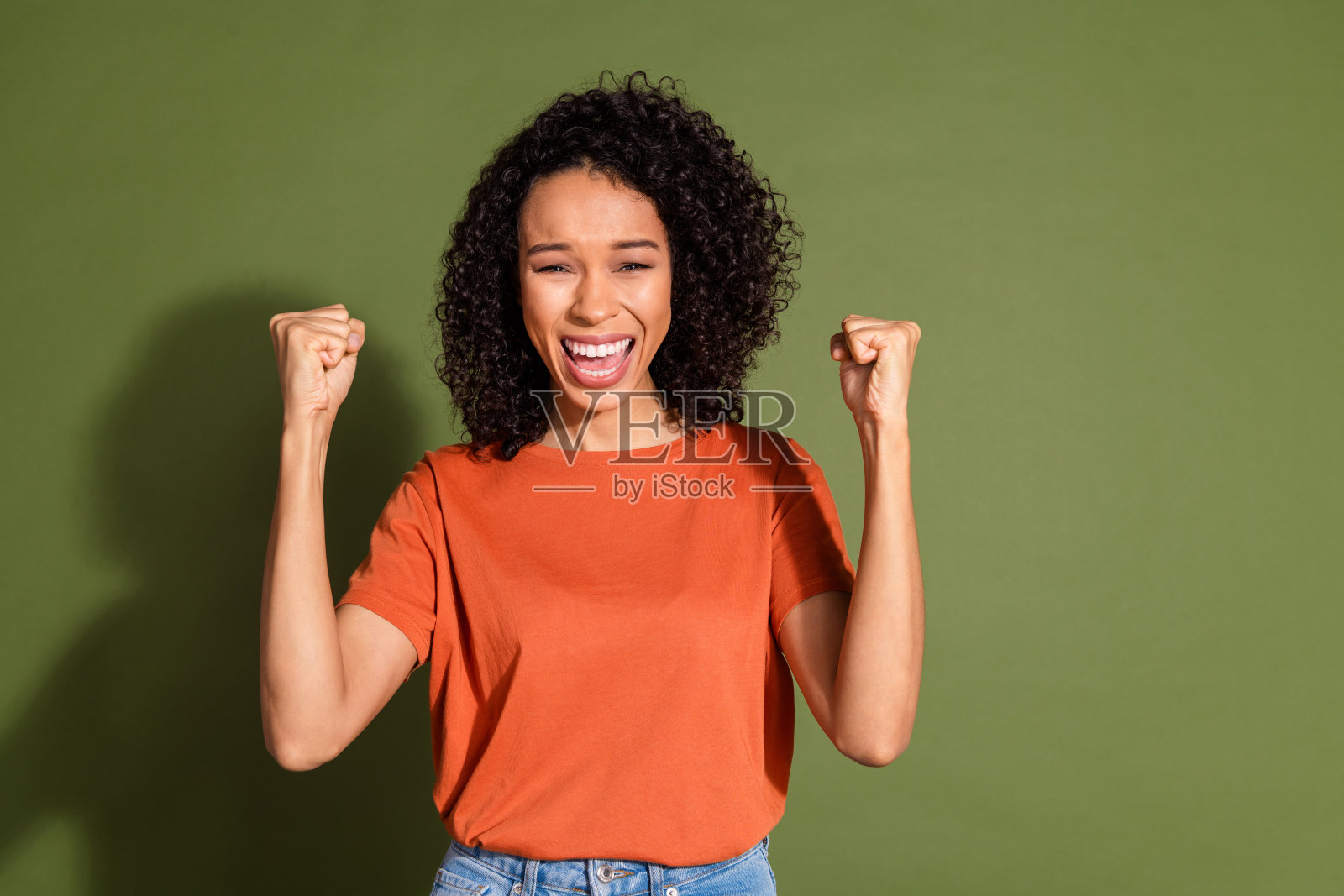 漂亮的年轻女孩的照片肖像赢得了举起拳头，穿着时髦的橙色服装孤立的卡其色背景照片摄影图片