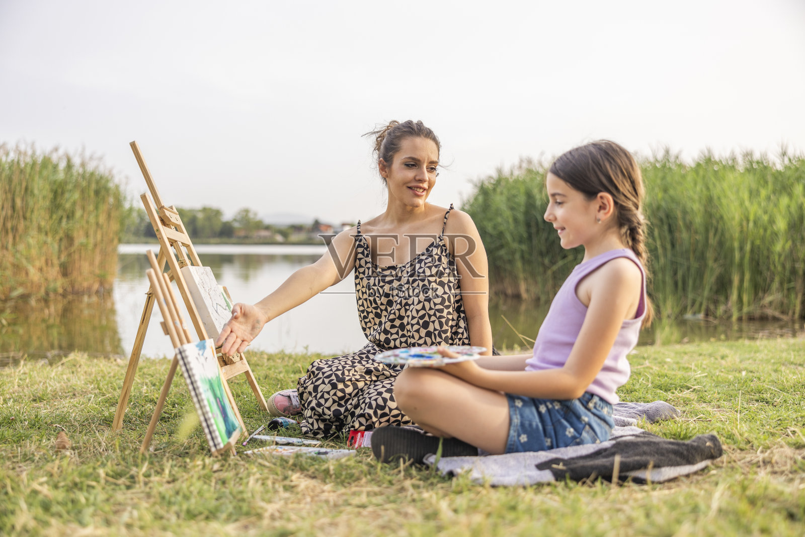老师在油漆上展示如何画一个孩子。他们正在湖边的户外上课。照片摄影图片