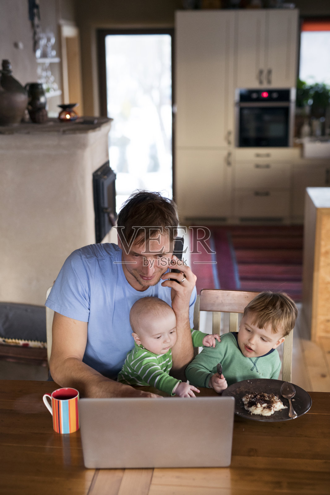 父亲坐在桌旁用笔记本电脑工作，两个儿子坐在他旁边。哥哥在吃粥。无条件的父母之爱，家庭时刻。照片摄影图片