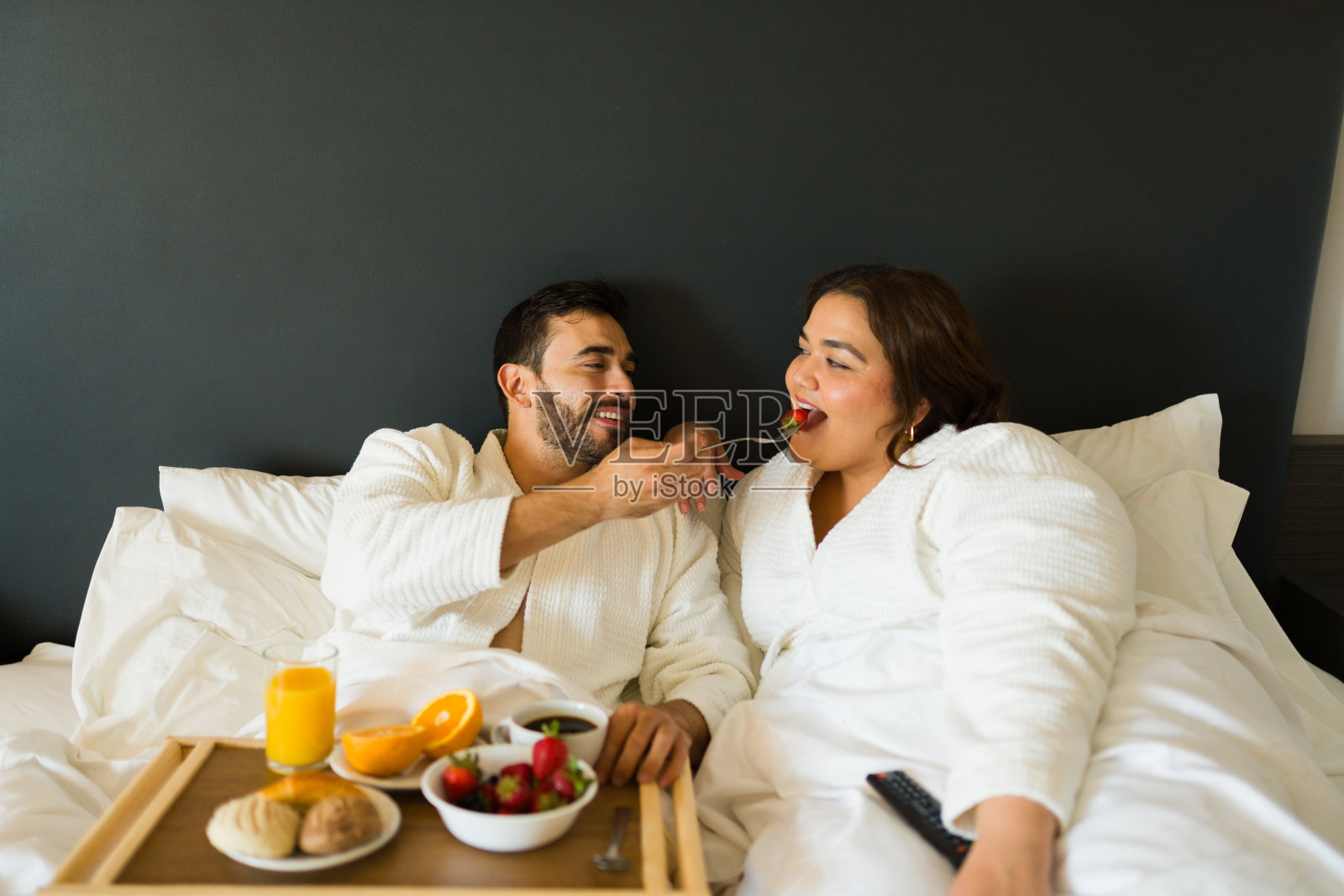 在酒店床上的浪漫早餐中，一个年轻人喂了一个草莓给他的女朋友照片摄影图片