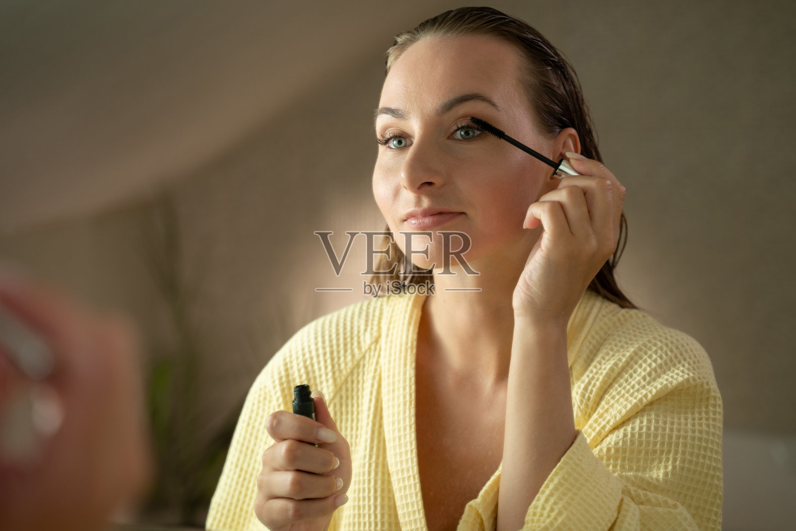 一位女士在浴室里用睫毛膏涂睫毛。她穿着一件淡黄色的长袍，正对着镜子。照片摄影图片