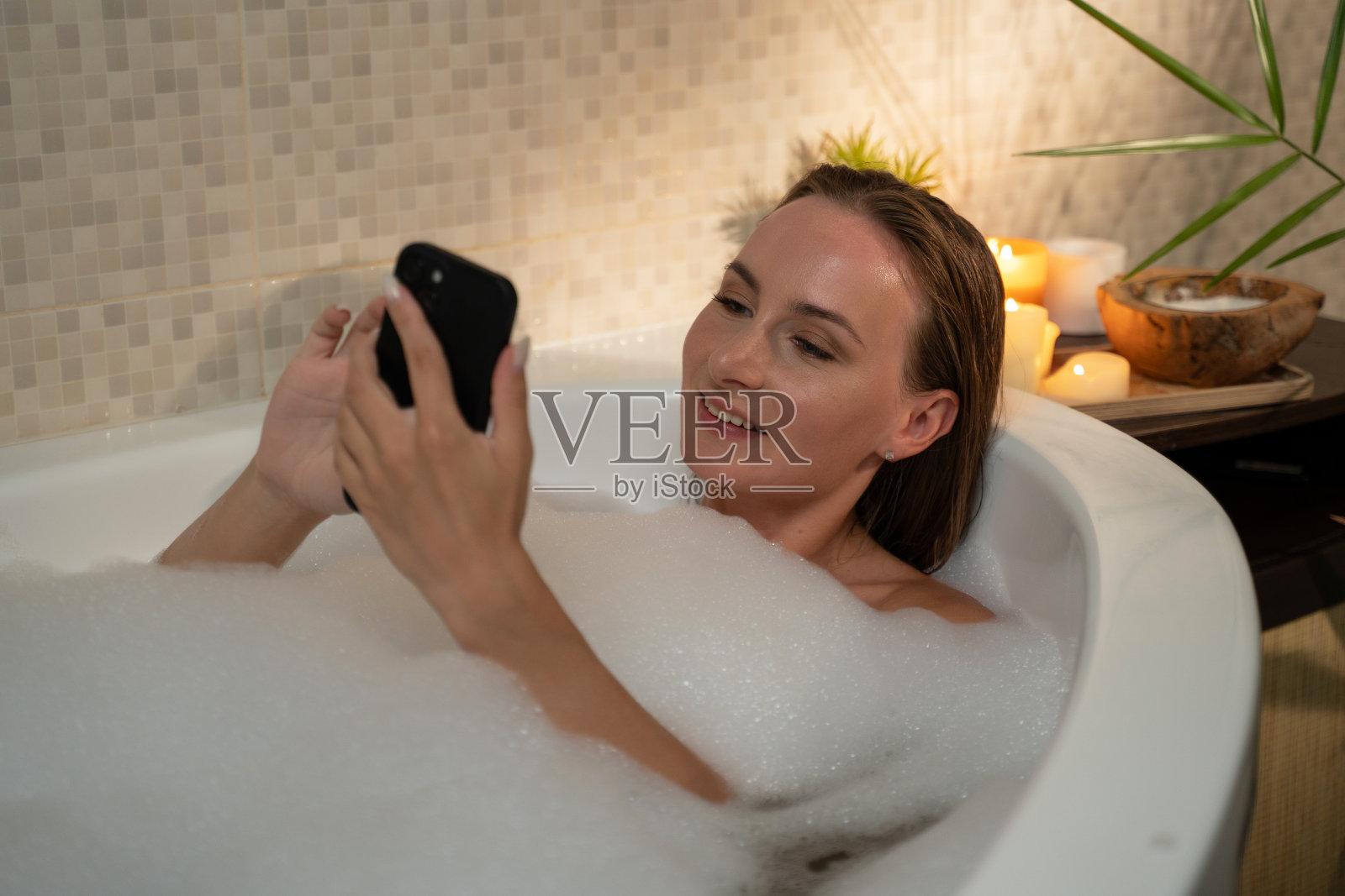 晚上在泡泡浴中放松的女人。她正在用智能手机上网或看视频。浴室里点着蜡烛，背景里有一株植物。照片摄影图片