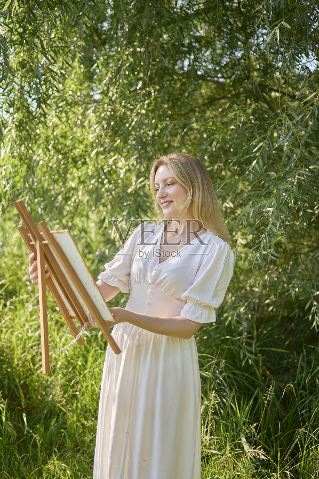 穿着白色连衣裙的年轻漂亮的艺术家站在河岸的树中间照片摄影图片