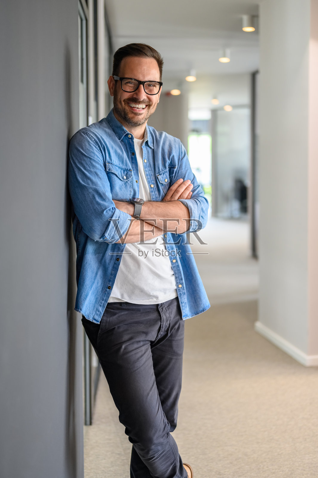 自信的商人，戴着眼镜，双臂交叉，靠在现代办公室的墙上照片摄影图片