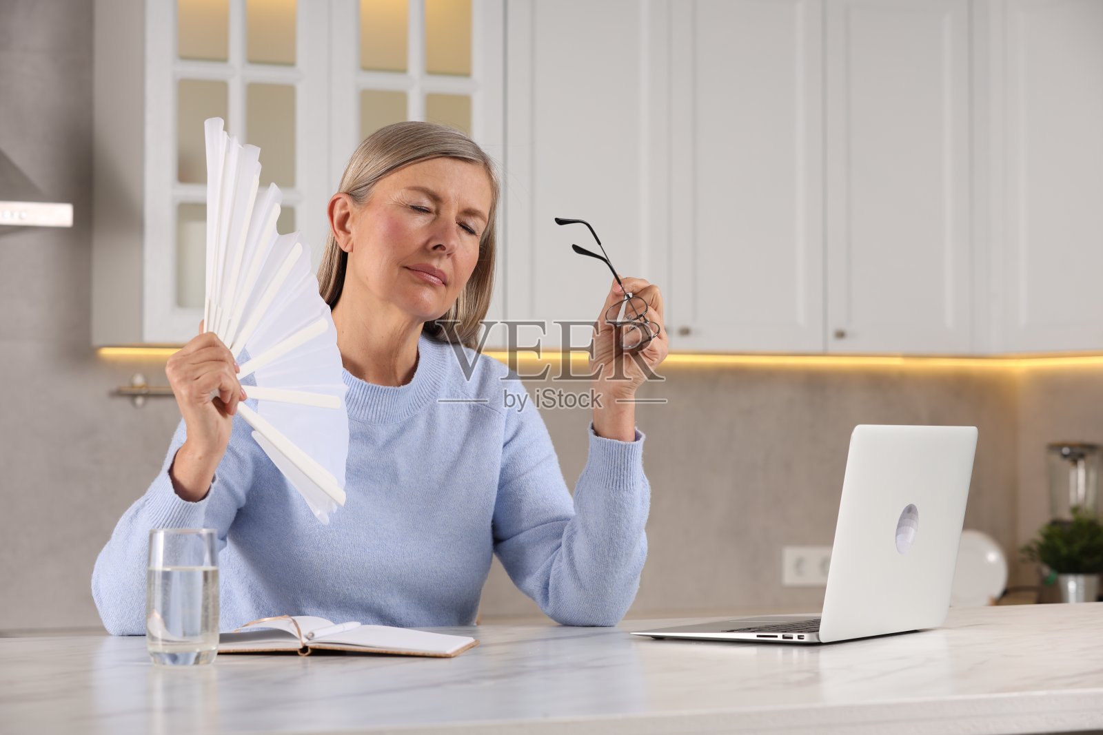 更年期。在厨房的桌子上，女人在潮热时挥舞着风扇给自己降温照片摄影图片
