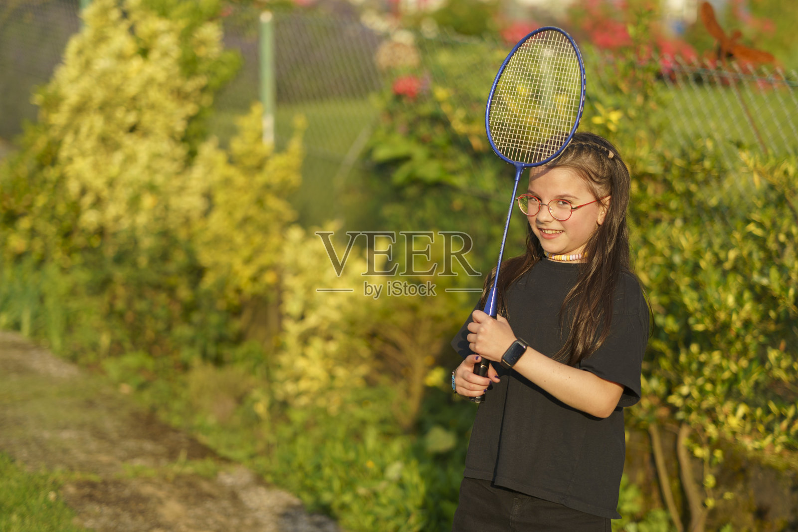 在一个阳光明媚的夏日，一个小女孩戴着眼镜，穿着黑色t恤和短裤，在她的后院打羽毛球。照片摄影图片