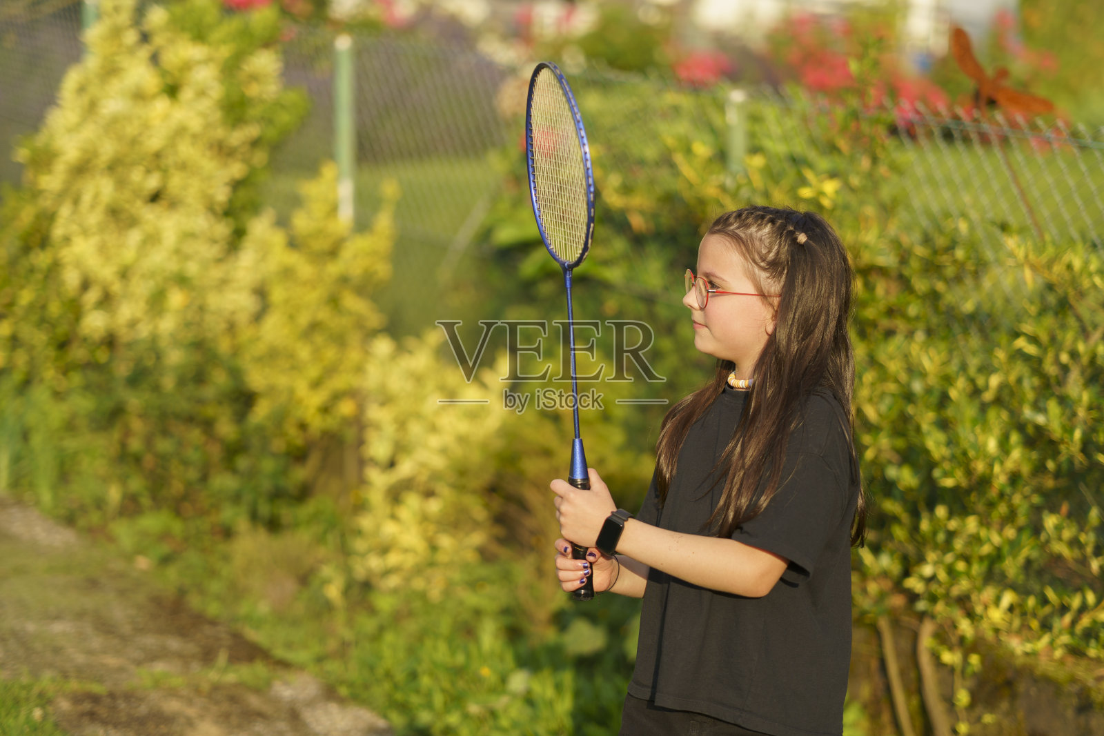 在一个阳光明媚的夏日，一个小女孩戴着眼镜，穿着黑色t恤和短裤，在她的后院打羽毛球。照片摄影图片