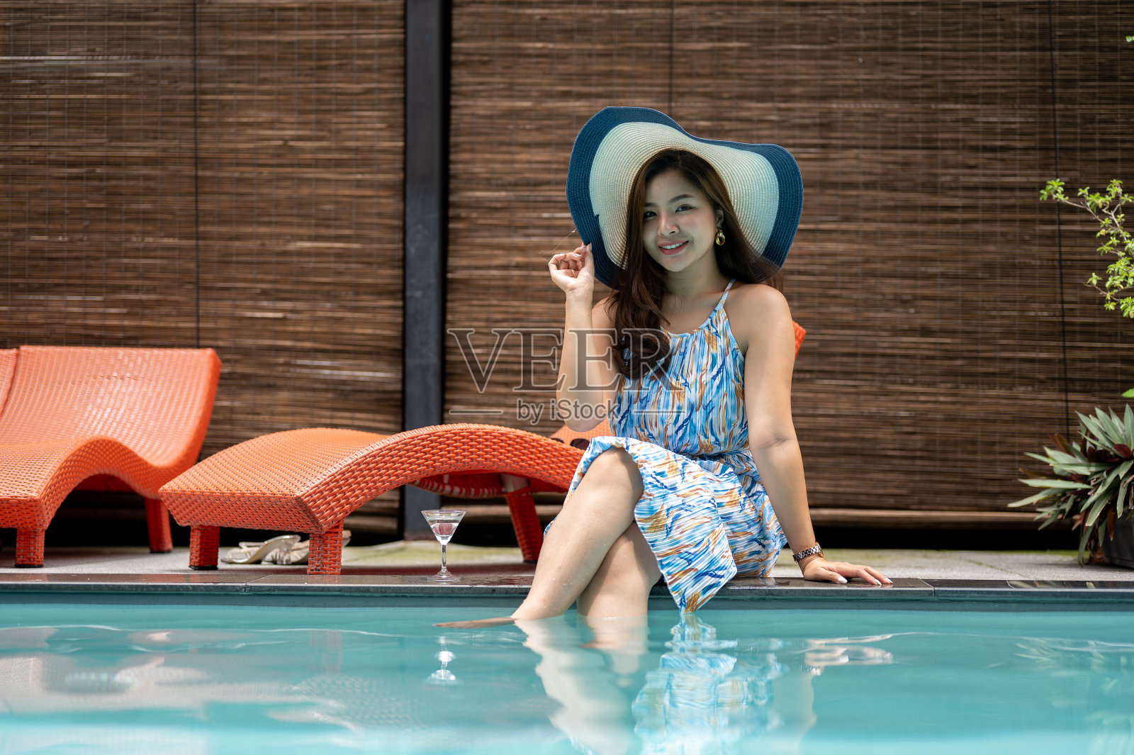 一个优雅、快乐的亚洲女人，穿着漂亮的夏装，头戴沙滩帽，坐在泳池边。照片摄影图片