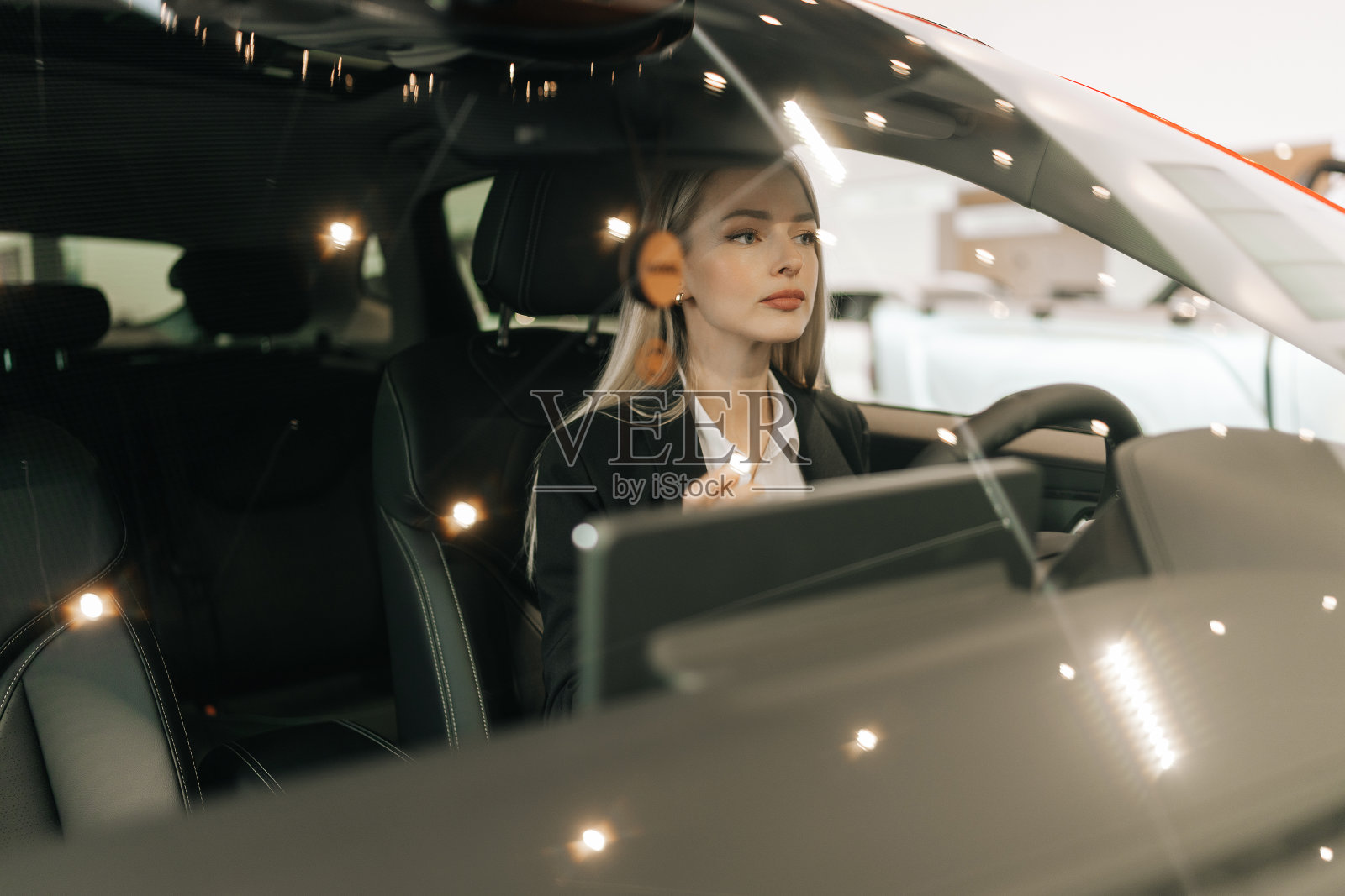 通过挡风玻璃拍摄到坐在车内驾驶员座位上的漂亮女顾客，在经销商处选择汽车试驾。女买家触摸方向盘照片摄影图片