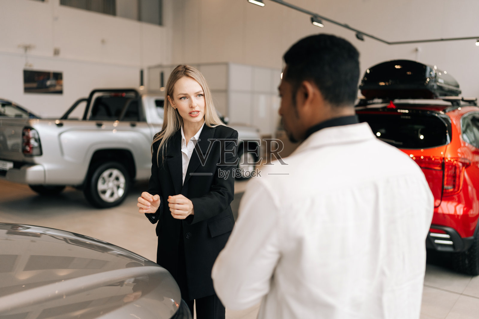 一个不知名的黑人买家的后视图，向专业的女性汽车经销商出售新车，解释其特征。穿西装的金发女售货员帮助挑选新车照片摄影图片