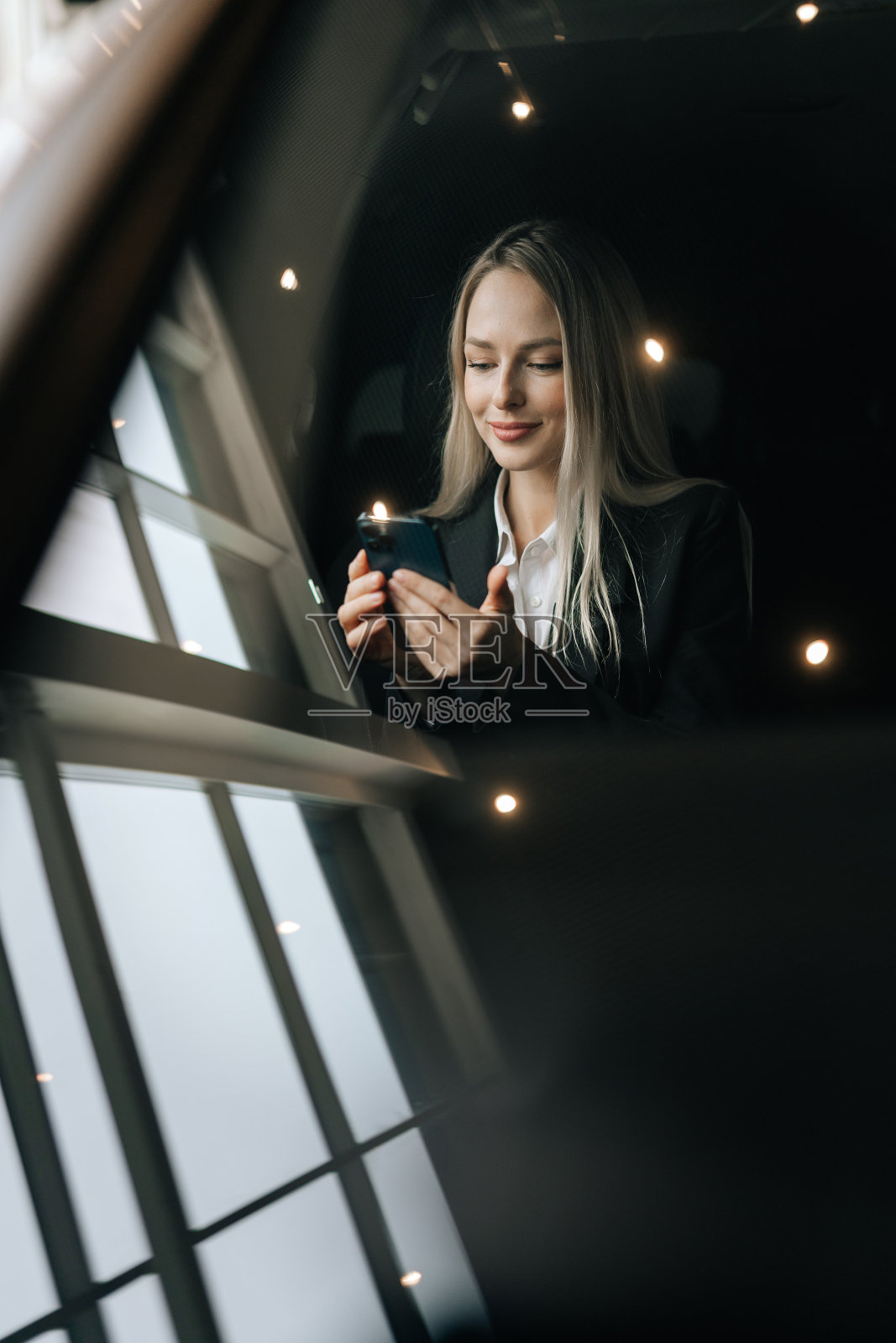 透过挡风玻璃的垂直照片，看到坐在汽车副驾驶座位上用智能手机阅读好消息的金发女郎。漂亮的年轻女子在浏览手机照片摄影图片