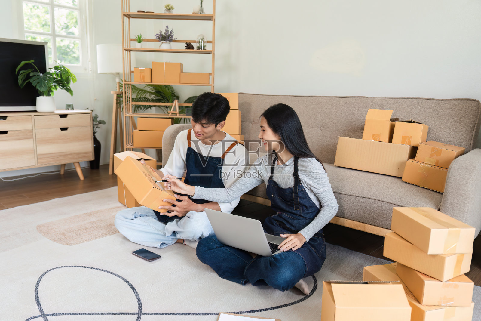年轻夫妇在家网上销售产品，周围都是纸板箱，使用笔记本电脑和智能手机照片摄影图片