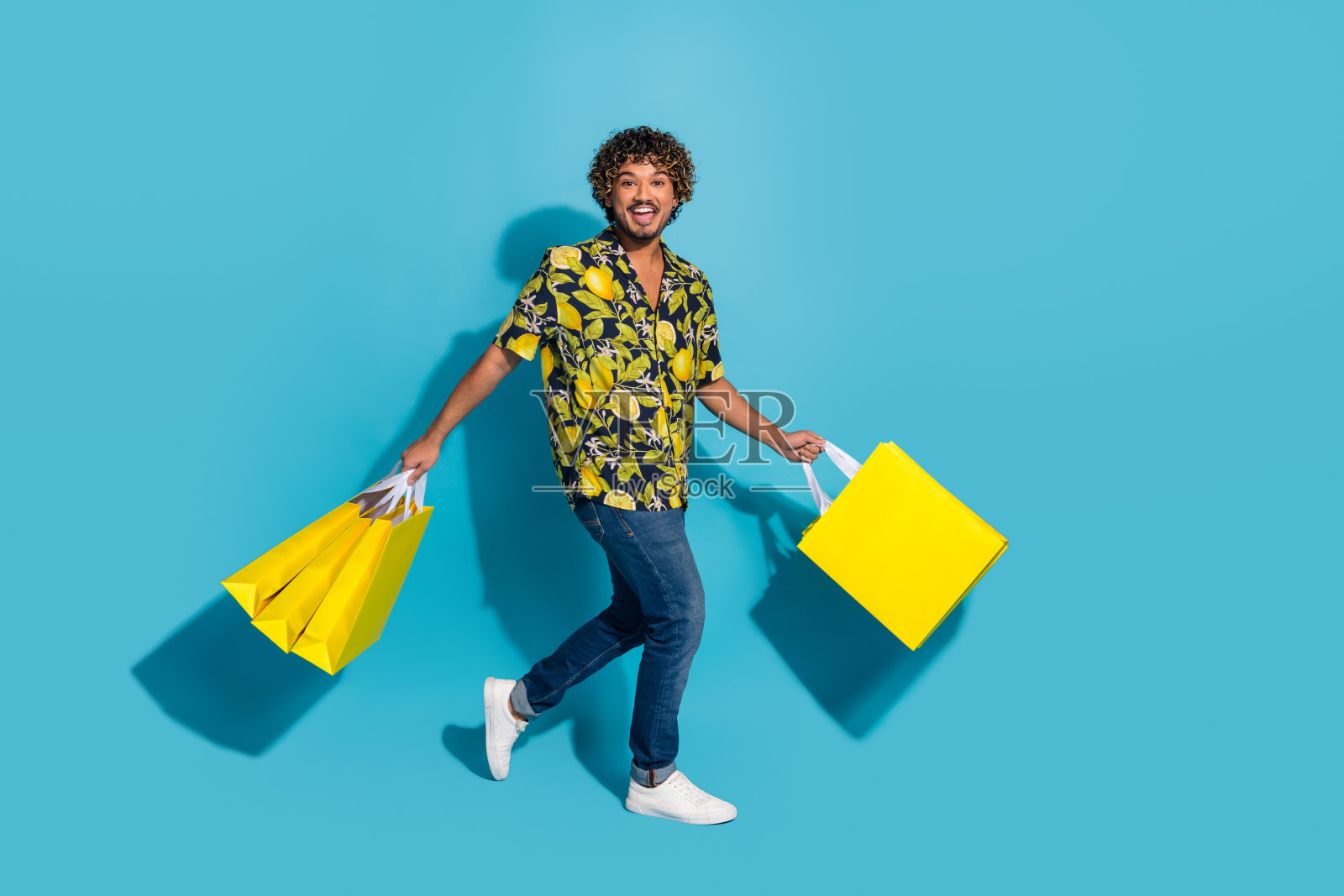 积极的男人穿着印花衬衫牛仔裤，拿着购物袋走到空旷的蓝色背景孤立的照片照片摄影图片