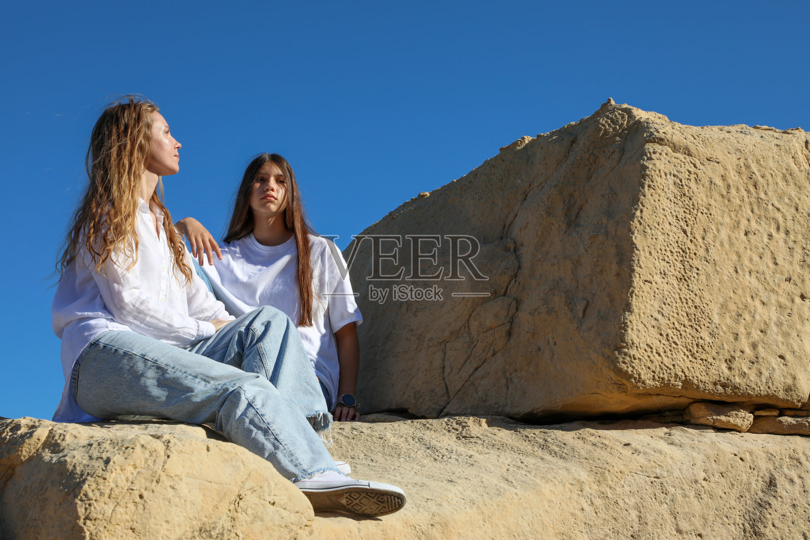 晴朗的蓝天下，一对母亲和十几岁的女儿坐在巨大的岩石上，她们都留着长发，穿着牛仔裤和白衬衫。照片摄影图片