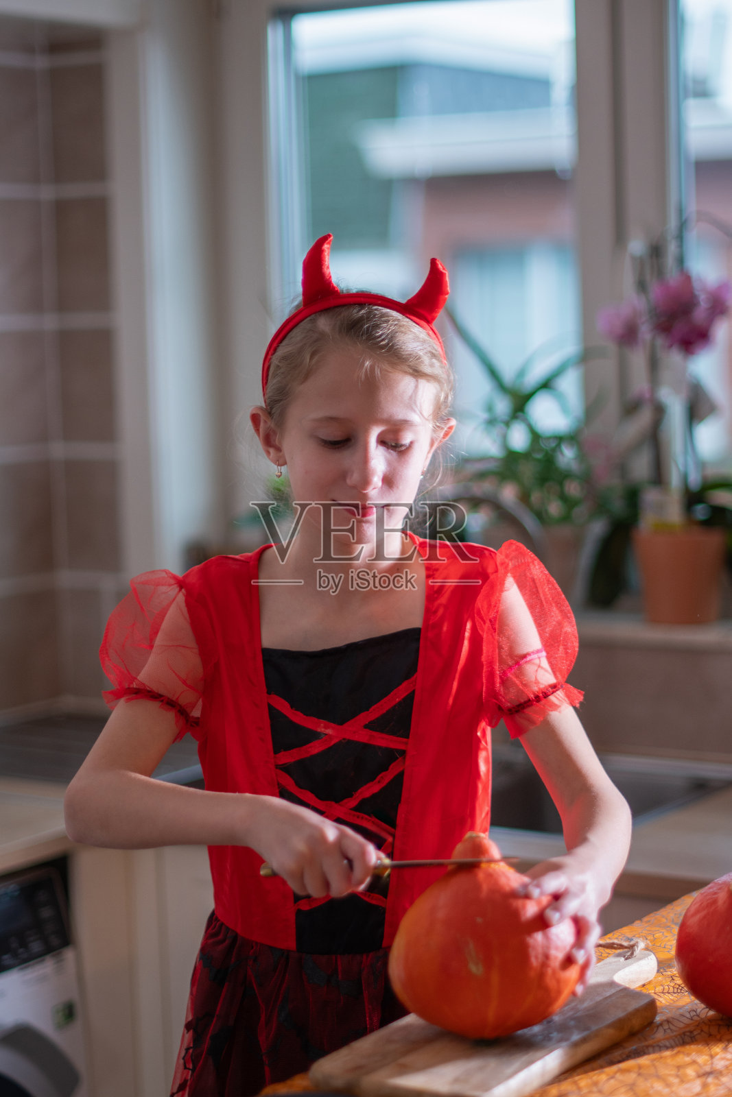 在黑暗的房间里，一个白人小女孩穿着万圣节的魔鬼服装，用一把锋利的刀切南瓜，脸上带着微笑照片摄影图片