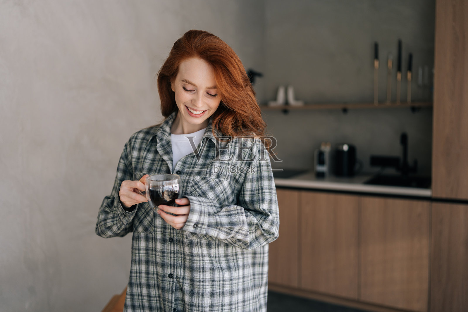 一个阳光明媚的早晨，站在厨房里，笑着拿着杯子喝咖啡的年轻女子的肖像。快乐微笑的女性独自在咖啡因休息时放松。照片摄影图片