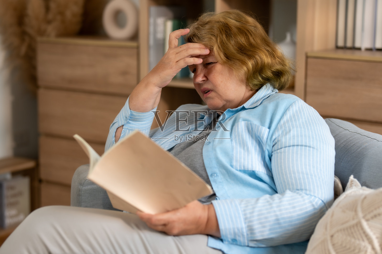 一位老妇人坐在舒适的客厅里，一边看书，一边感到不适，这凸显了她对家庭护理和支持的需求。照片摄影图片