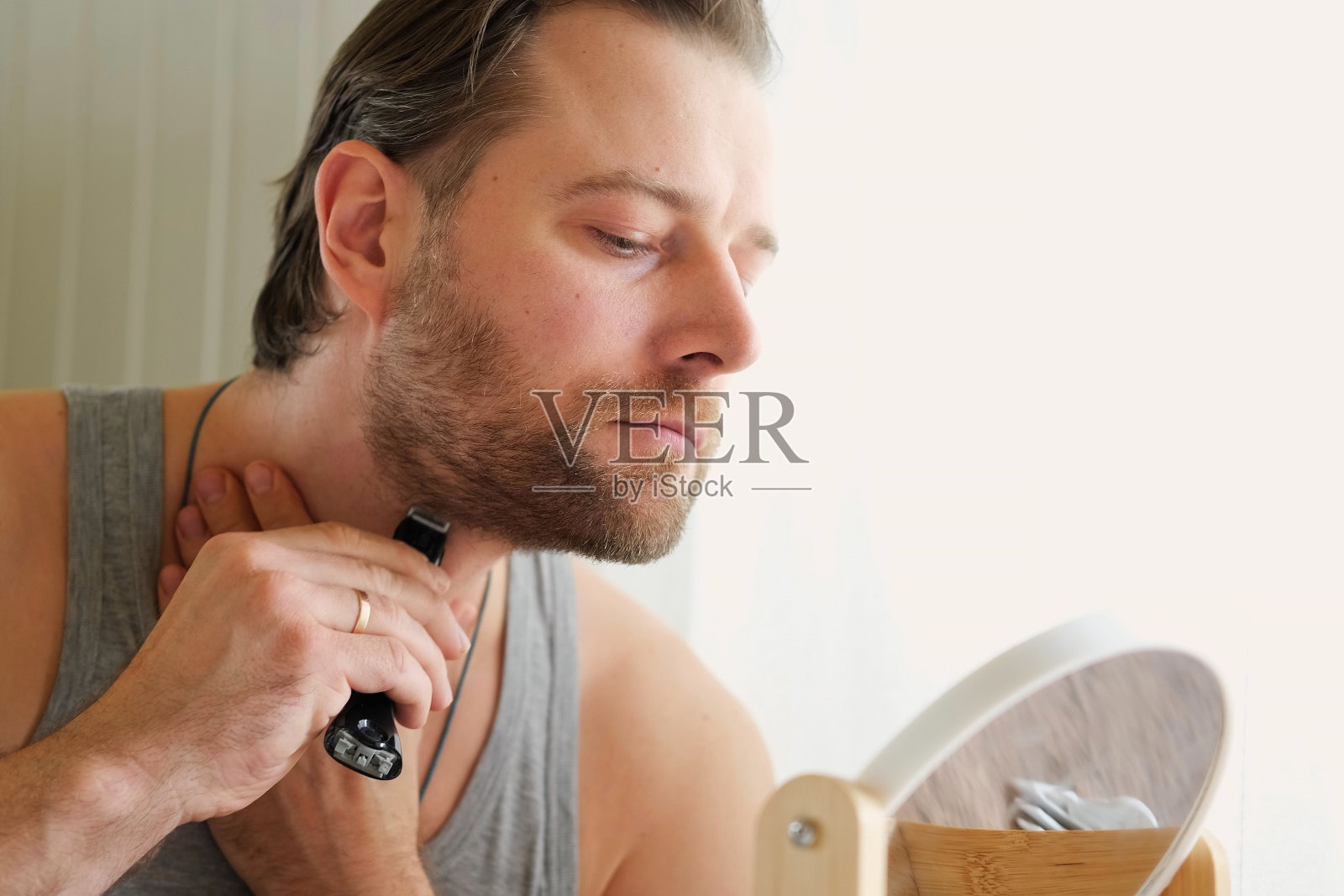 一个留着胡子的年轻人拿着电动剃须刀在浴室里刮胡子的倒影。帅哥坐在镜子前。30多岁男性，用修剪器修剪胡须。早上例行照片摄影图片