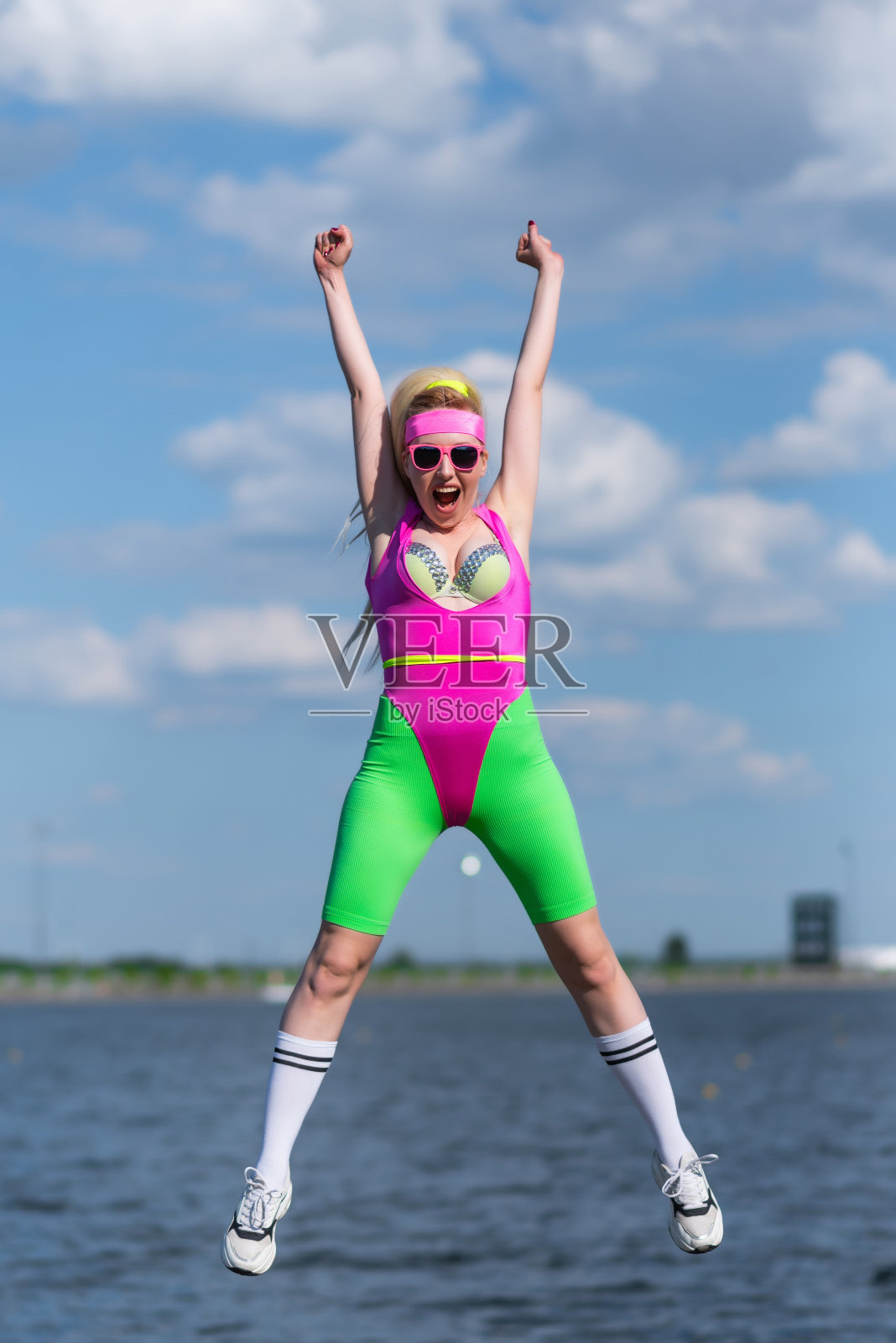 年轻美丽的金发女子穿着运动短裤，粉红色的紧身衣和粉红色的太阳镜在户外做运动。跳向天空。运动，积极的生活方式，运动训练，健康的生活方式照片摄影图片