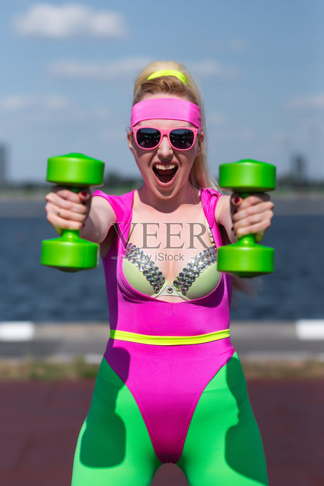 年轻美丽的金发女子穿着运动短裤，粉红色的紧身衣和粉红色的太阳镜在户外做运动。用哑铃做练习。运动，积极的生活方式，运动训练，健康照片摄影图片