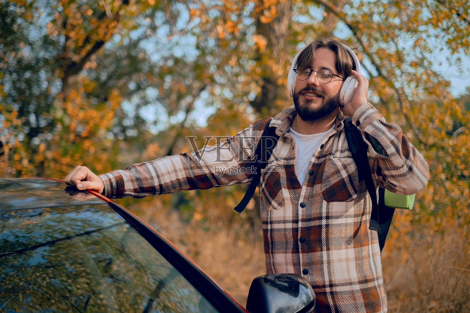 留着胡子的十几岁男孩，穿着衬衫和牛仔裤，坐在森林里，戴着耳机，听着放松和冥想的音乐，唱着音乐。照片摄影图片