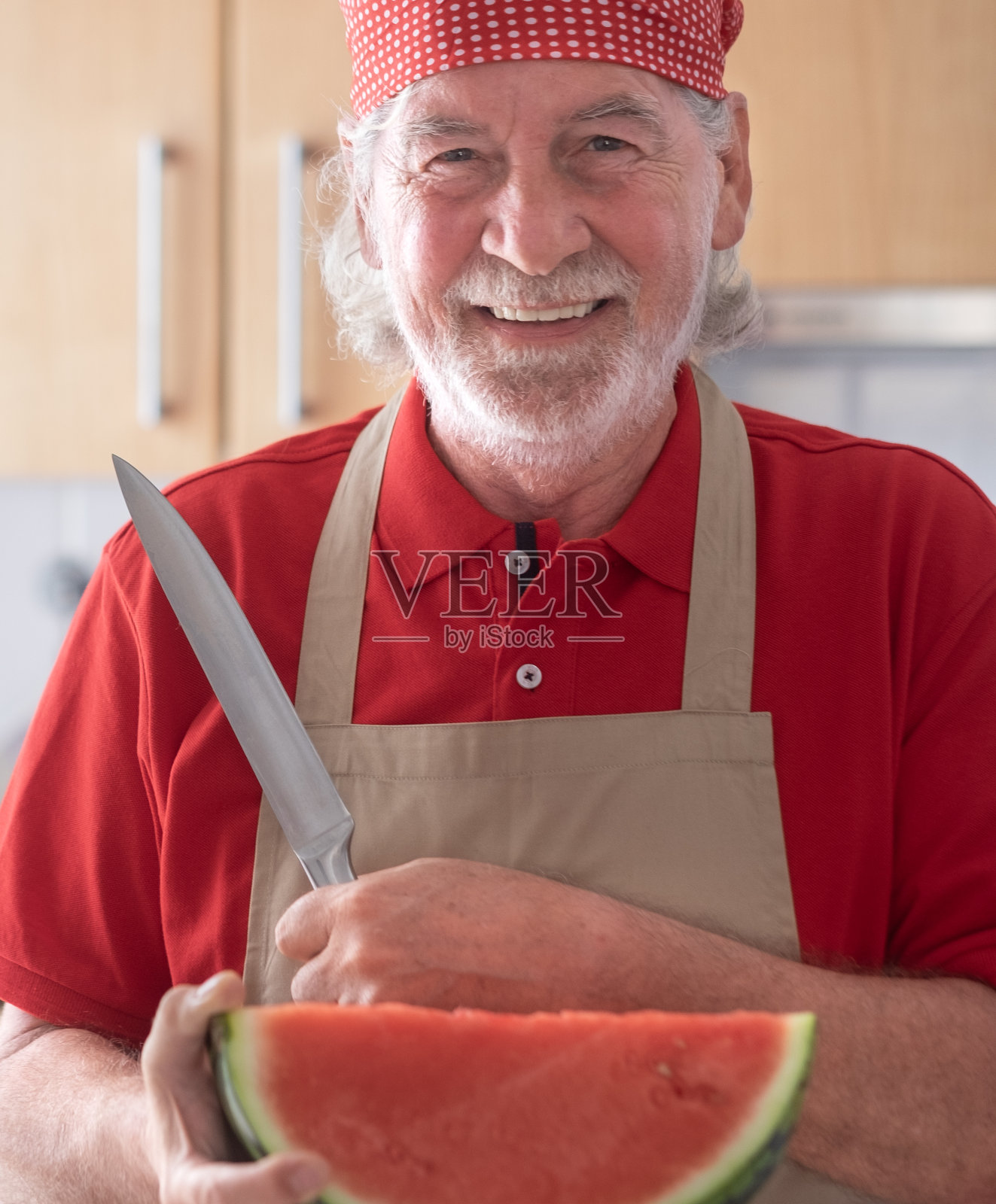 一位身穿红衬衫、戴着头巾的老人，脸上带着欢快的微笑，手里拿着一把大刀，手里拿着刚切成两半的西瓜。西瓜又红又多汁，健康又清爽的零食照片摄影图片