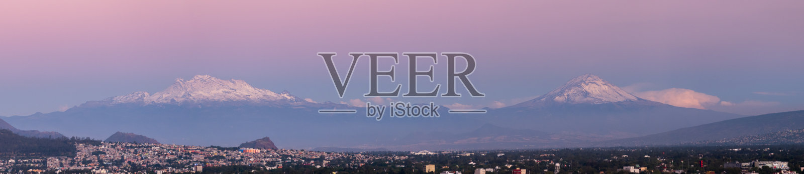 墨西哥城坐落在两座白雪皑皑的火山脚下，在柔和的粉红色黄昏的天空下。照片摄影图片