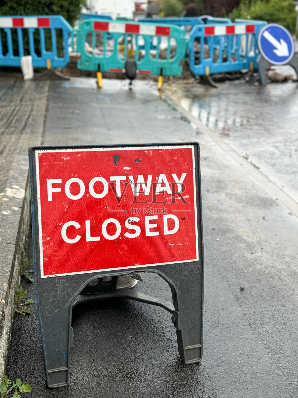 居民区柏油路面上的红白警告标志“道路关闭”的特写图像，带有沙袋平衡物的“A”框架标志，绿松石，塑料安全屏障，潮湿的天气条件，重点在前景照片摄影图片