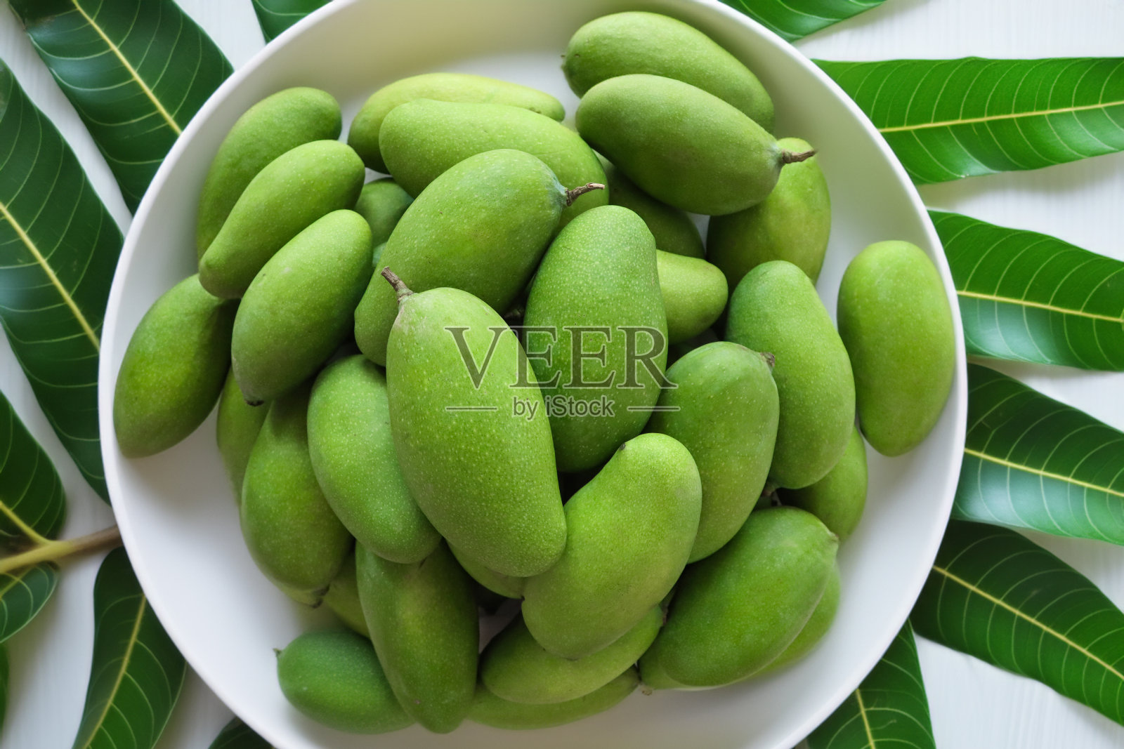 一堆完整的，生的，热带芒果果实堆在一个白色的盘子里，绿色的皮肤，闪亮的叶子，白色的背景，抬高的视角照片摄影图片