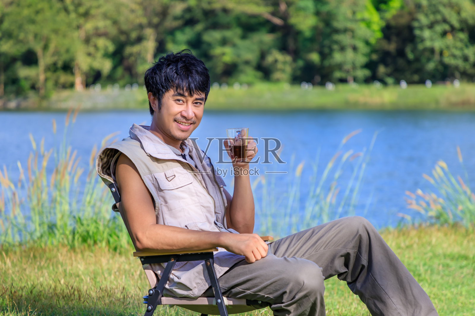 年轻的亚洲男性坐在椅子上，喝着咖啡，放松地举手，在湖边和山上露营。看着镜头。照片摄影图片