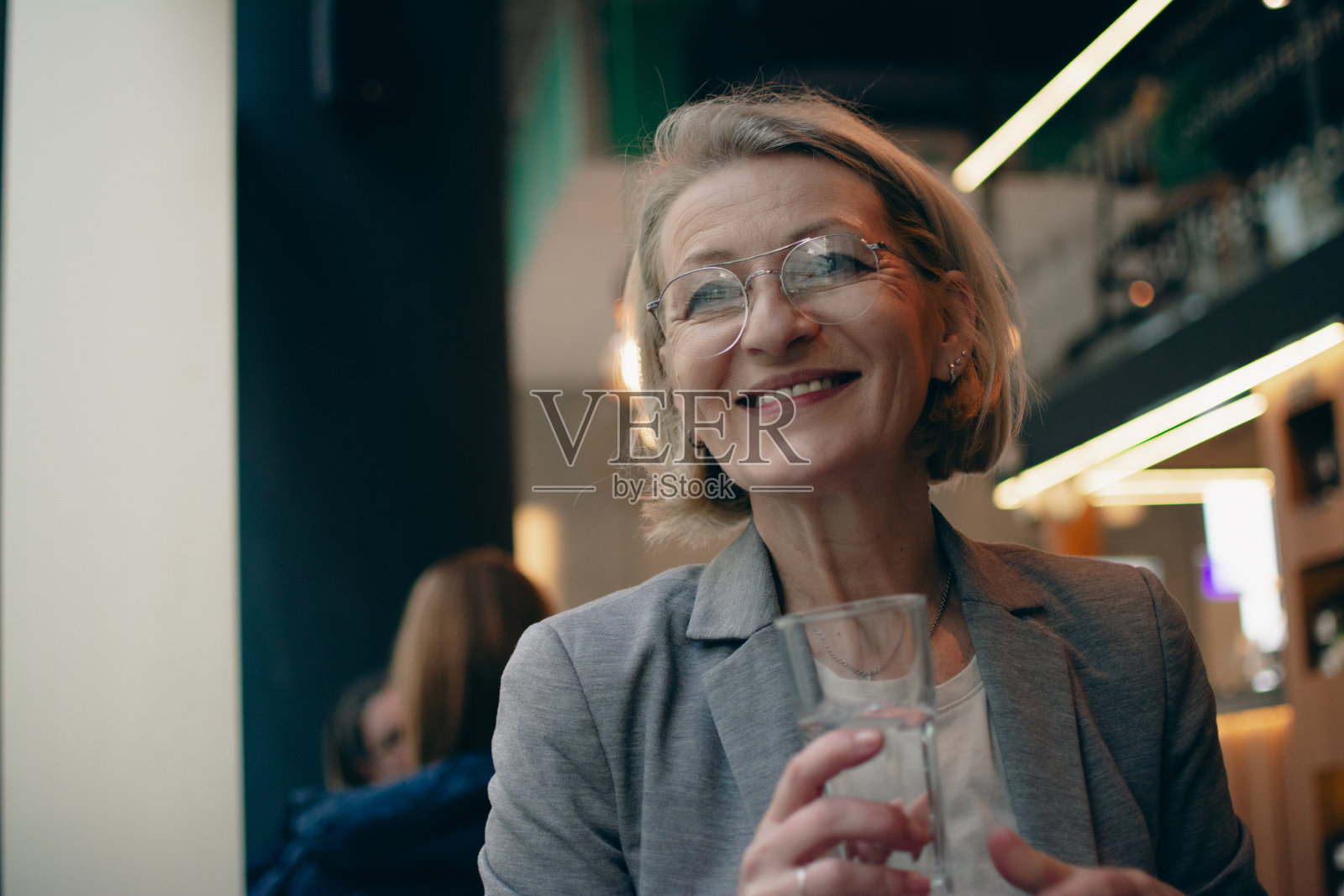 戴着眼镜的成熟金发女商人坐在自助餐厅里，一边若有所思地看着窗外，一边用杯子喝水照片摄影图片