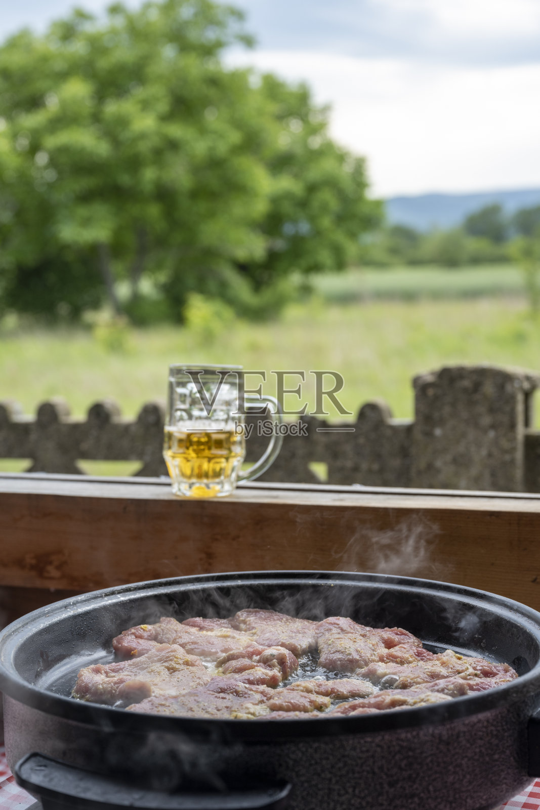 用特制的平底锅煎或炖猪肉、牛肉、小牛肉。乡村的夏日厨房。在背景中，你可以看到一杯啤酒，以及草地和森林的美丽景色。照片摄影图片