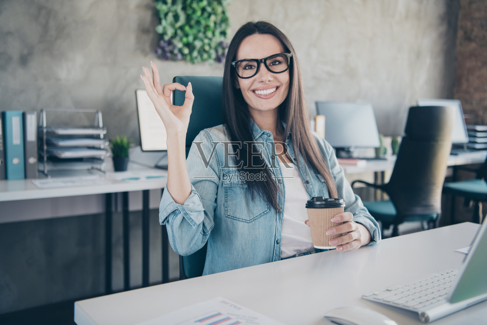 快乐迷人的女士助理的照片穿牛仔裤衬衫显示ok喝饮料在室内车间工作站阁楼照片摄影图片