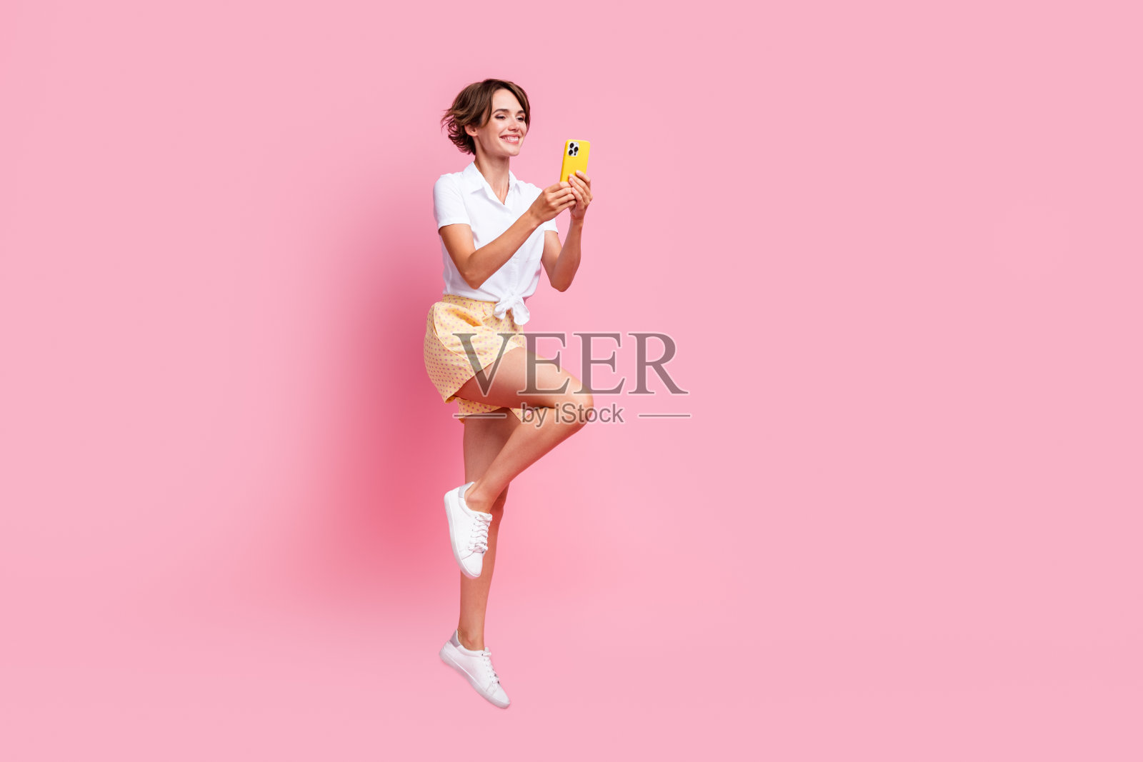 迷人的年轻女孩全身跳蹦床，同时使用她的全新iphone 15发布孤立的粉红色背景照片摄影图片