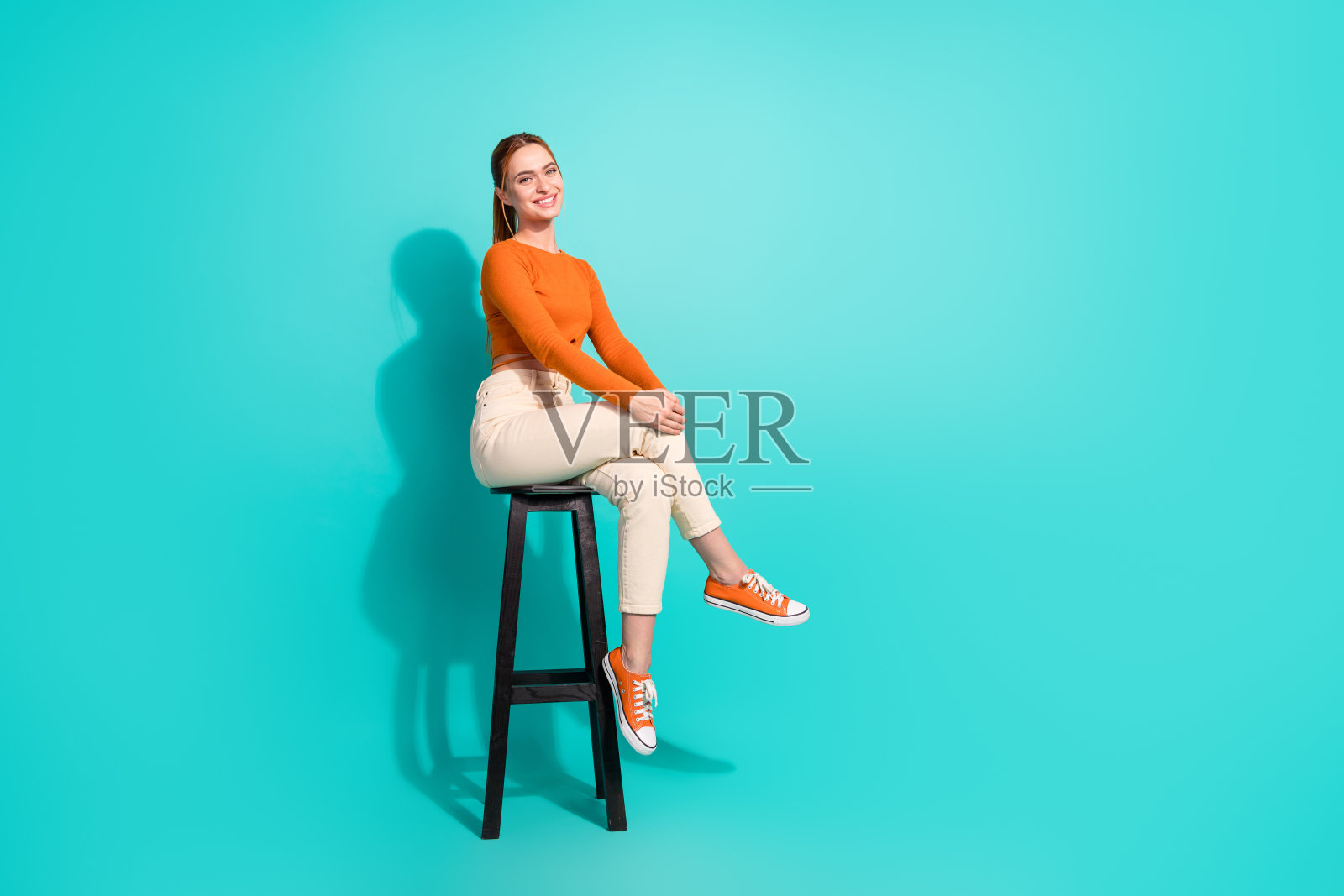 成功女性的全身照片，穿着露臀裤，坐在酒吧椅子上，双腿交叉，在绿松石色的背景上分开照片摄影图片