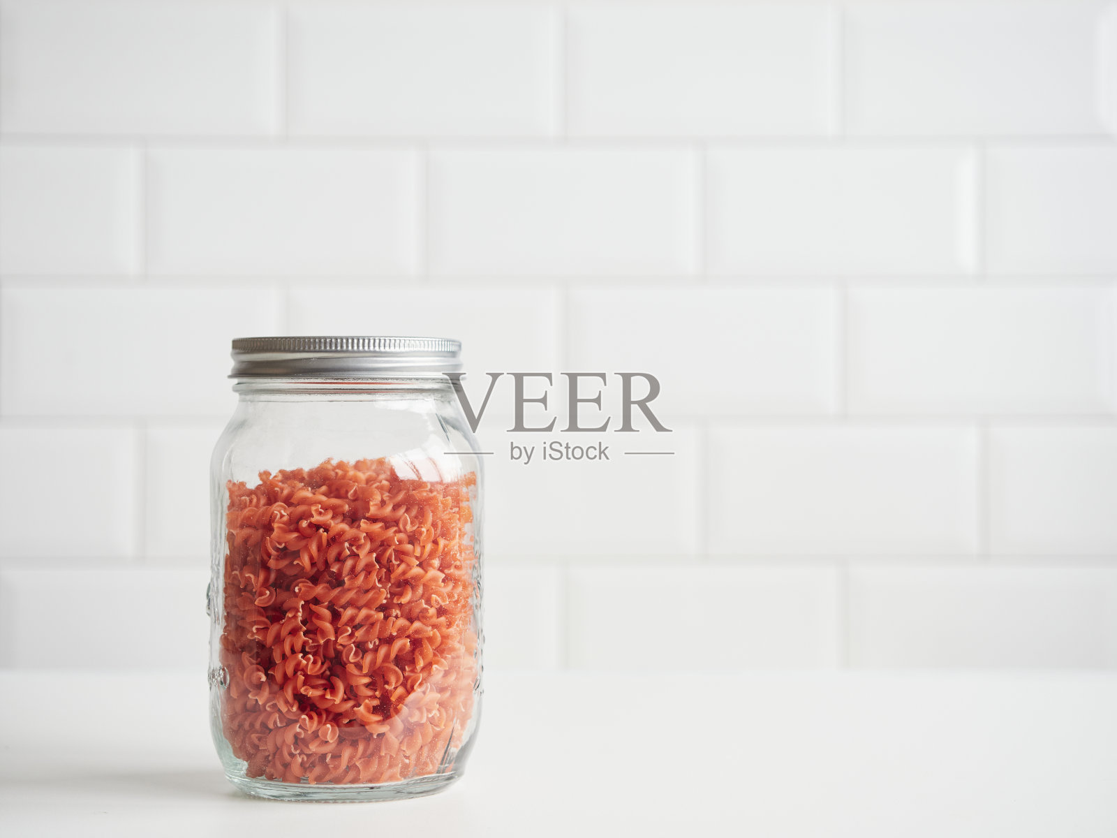 在厨房食品储藏室储存食物。干红扁豆意面，螺旋状，可重复使用的有盖玻璃罐中。前视图上的白色瓷砖模糊的背景。照片摄影图片