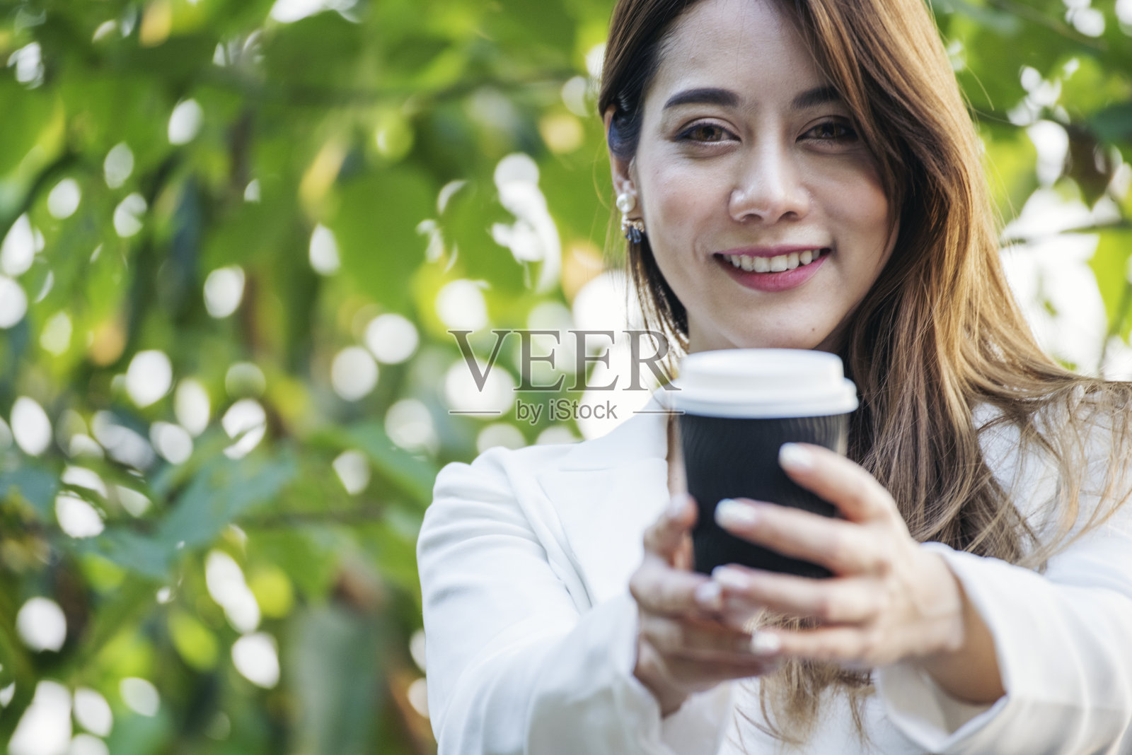 在绿色公园里喝咖啡的女人手拿热的一次性杯子。快乐放松美丽的亚洲女人微笑的脸站在户外花园。年轻女性享受大自然的早晨自由的生活方式。照片摄影图片