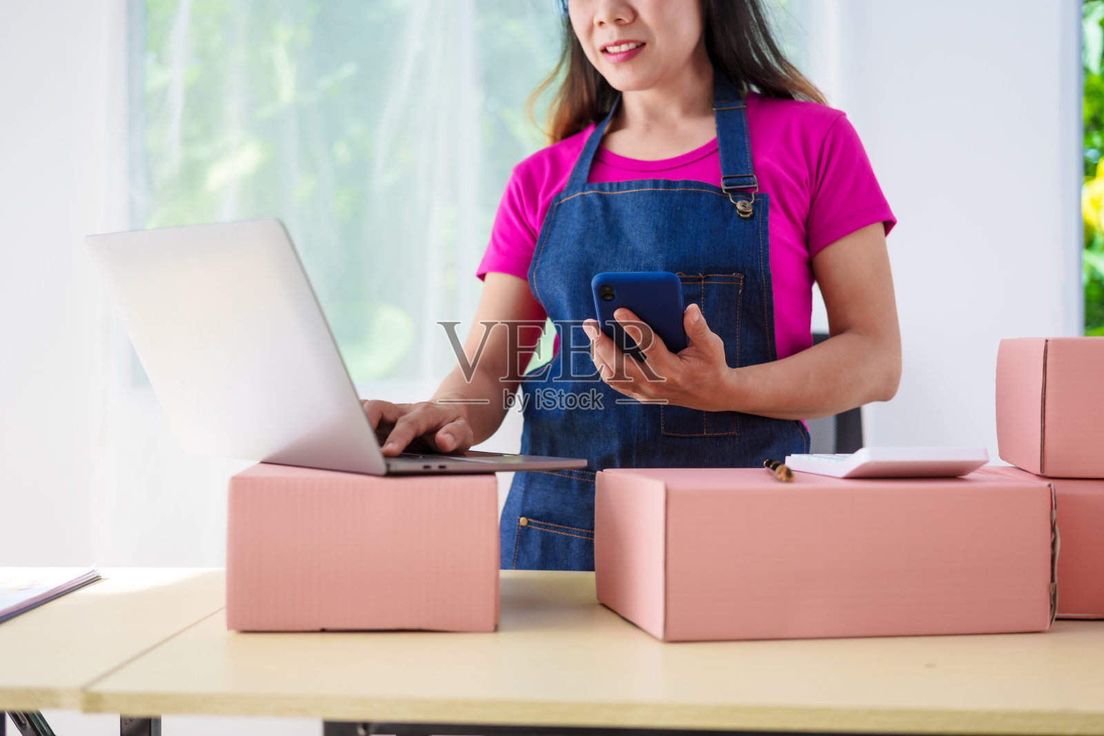 一个穿着工作服的女人在网上销售产品，检查包裹，接受客户订单，在办公桌前工作，用粉红色的盒子包装物品，准备运送给客户。照片摄影图片