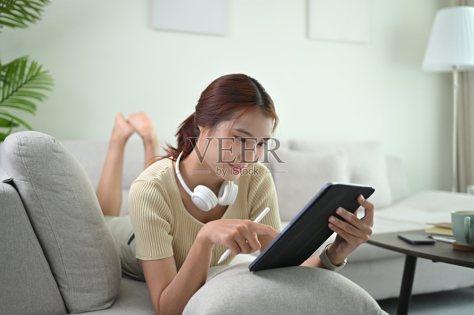 漂亮的年轻女子穿着休闲服躺在沙发上，在电子平板电脑上查看社交媒体照片摄影图片