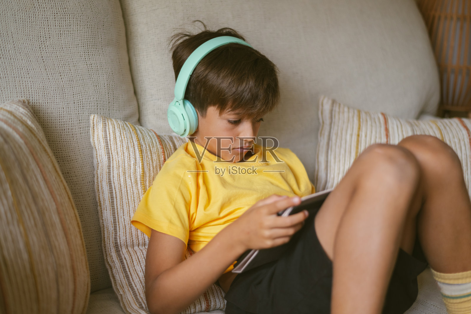 男孩坐在沙发上，戴着耳机盯着他的平板电脑屏幕照片摄影图片