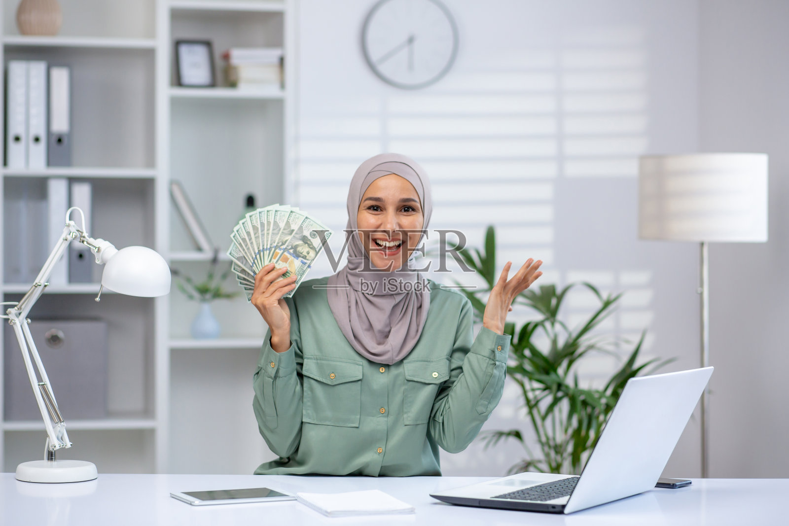 在现代办公环境中，微笑的女人拿着现金，用笔记本电脑工作照片摄影图片